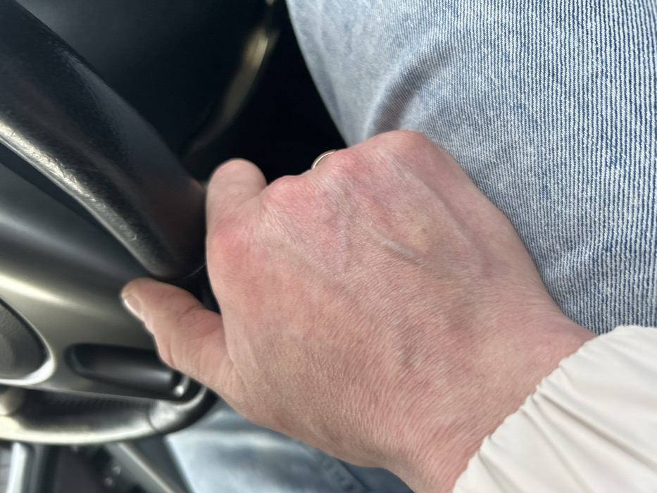 A close-up image depicts an adult's left hand resting on a vehicle's steering wheel, originating from the city of Ir'yak, Russia. The hand, exhibiting fair skin with visible veins and slightly reddened knuckles, is positioned with the thumb and index finger gripping the upper-right portion of a dark-colored, possibly black, steering wheel rim. A metallic ring is present on the ring finger. The steering wheel features a lighter, possibly silver-colored, inner spoke or trim. Below the hand, the individual is wearing light blue denim jeans. A portion of a light-colored, likely white or cream, long sleeve is visible on the left side of the frame, near the wrist area. The scene is illuminated from an unseen source, casting subtle shadows.