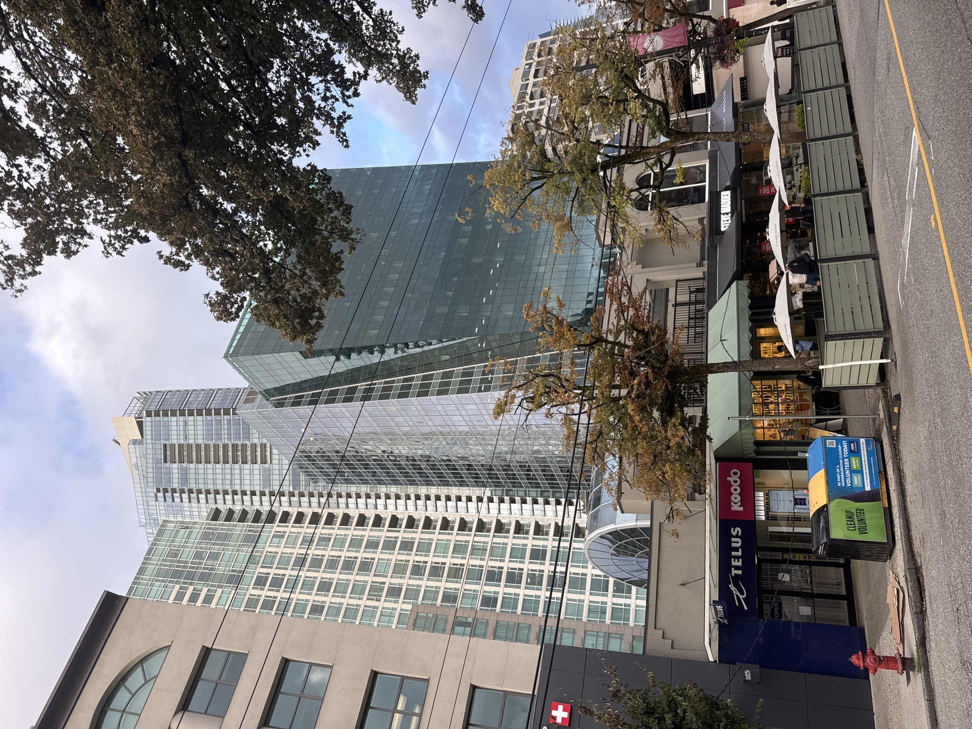 This is a street-level, medium-angle, eye-level shot of a city scene, likely in Vancouver, Canada. The image is dominated by modern, tall buildings with glass facades, interspersed with some older, more traditional architecture at street level. In the foreground, a sidewalk area features outdoor seating with umbrellas, suggesting a commercial district with restaurants or cafes.
Several businesses are visible: a Telus and Koodo store, a "Steve Madden" shoe store, and a "Ladurée" establishment. A bright red fire hydrant stands near the Telus store. A large blue and yellow bin, marked "CLEANUP VOLUNTEER," is placed on the sidewalk.
The sky is overcast with a mix of grey clouds and some lighter patches. Trees with autumn foliage are present, adding a natural element to the urban landscape. There are a few people visible on the sidewalk, some walking and others seated at the outdoor tables, but their faces are not clearly discernible. Power lines and streetlights are also visible, indicating a typical urban street environment. The overall impression is a bustling, modern city street with a blend of architectural styles and commercial activity.