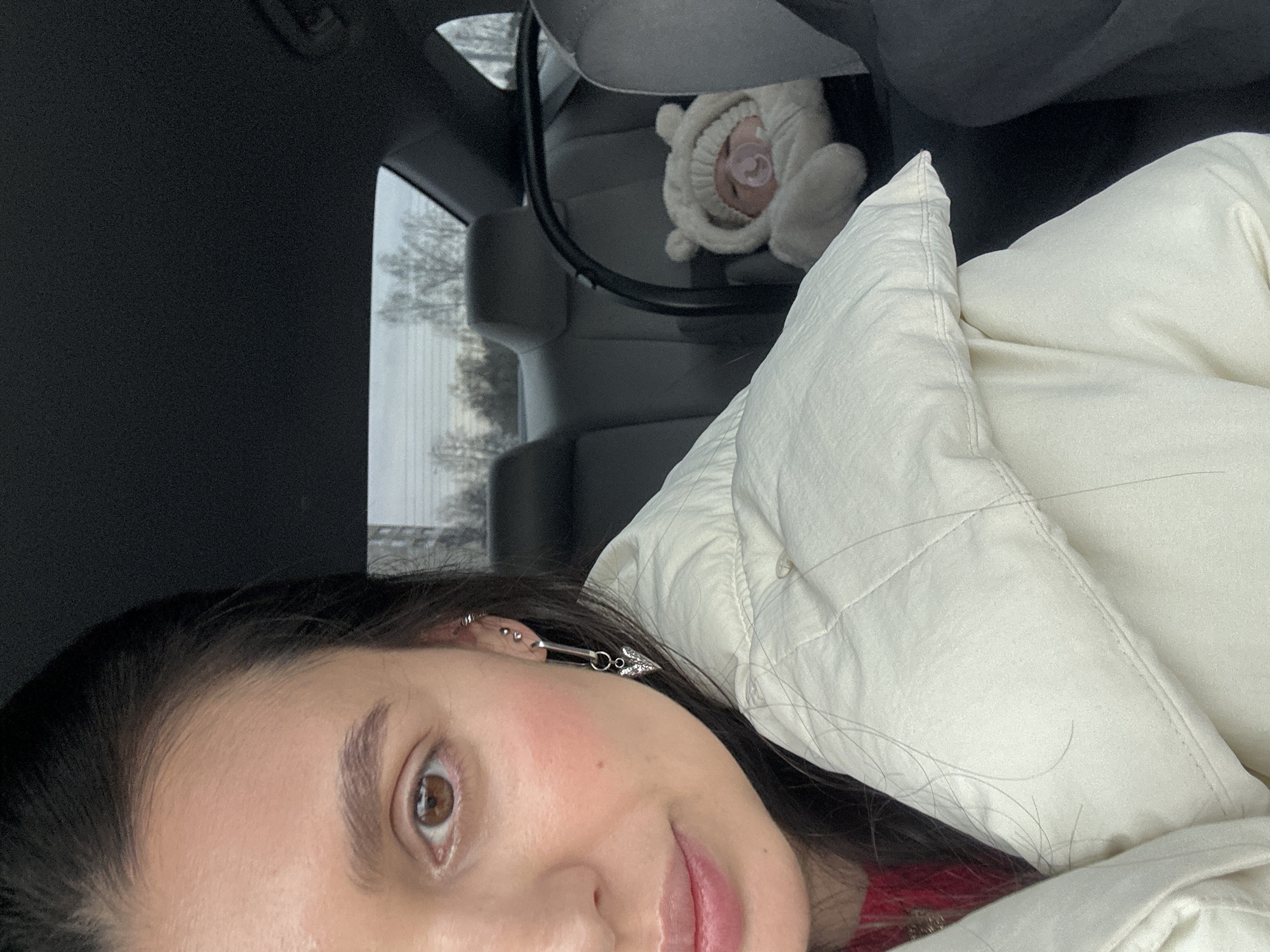 The image appears to be a selfie taken inside a vehicle, likely a car, with a baby in a car seat visible in the background. The woman taking the selfie is wearing a white quilted jacket and has dark hair with piercings in her ear. The baby is wearing a white hat with bear ears and has a pacifier in its mouth. The background shows a glimpse of a gray car interior and a window with a view of trees outside. It is not possible to determine the location in Chaikovsky, Russia from the image.