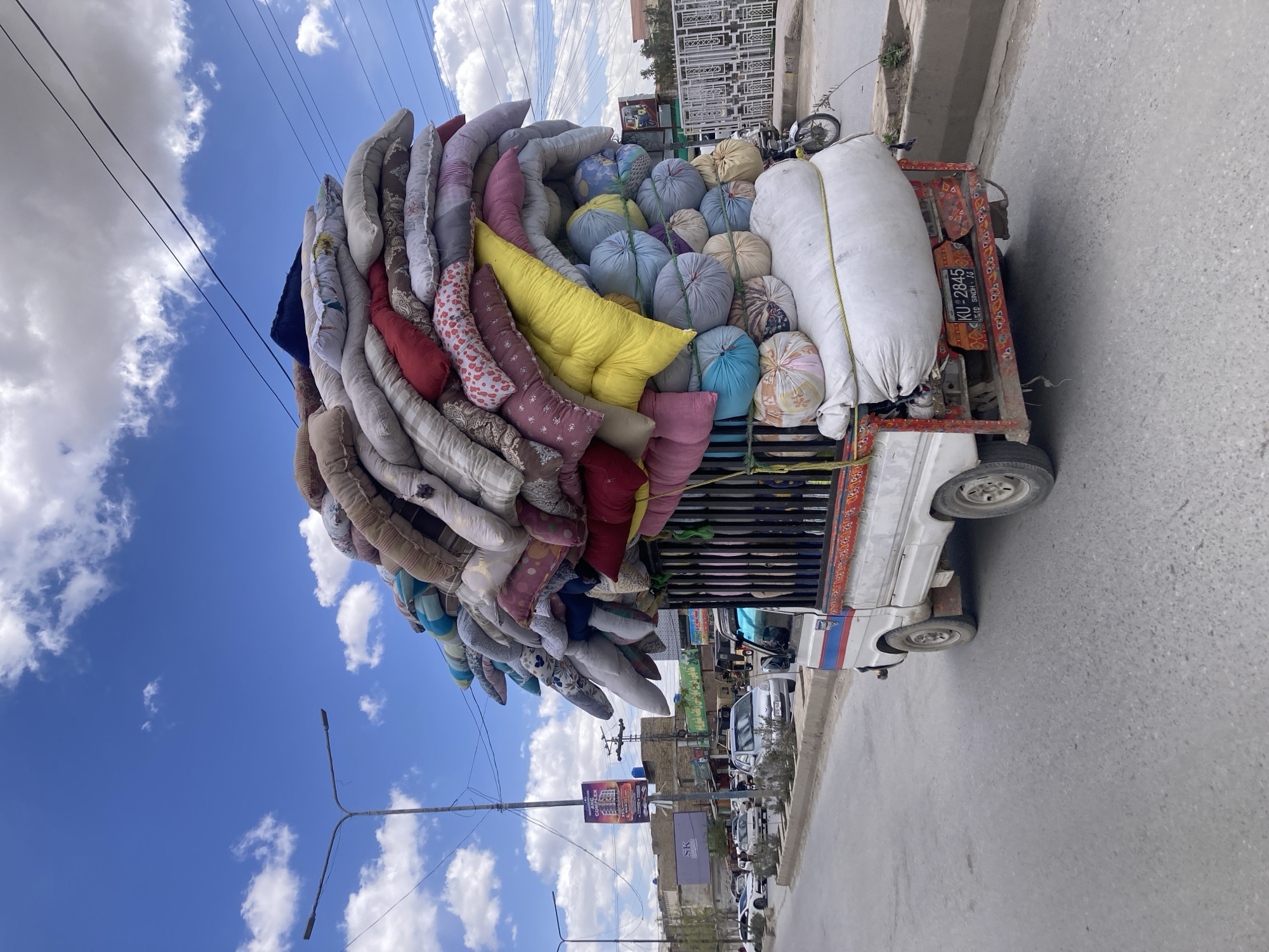 A motor vehicle is heavily loaded with fabric and bedding material, likely for transport or sale, on an urban street in Quetta City Tehsil, Pakistan. The vehicle, identified by the number plate "KU-2845 SINDH-73," is oriented vertically in the frame with the load reaching high above the vehicle. The load is a mix of pillows, bedding rolls, and sacks, all secured to the vehicle's cargo area. The street has a sidewalk and buildings on either side. Utility poles with overhead lines are present along the street. The sky above is blue with scattered clouds. Other vehicles and pedestrians are visible in the background.