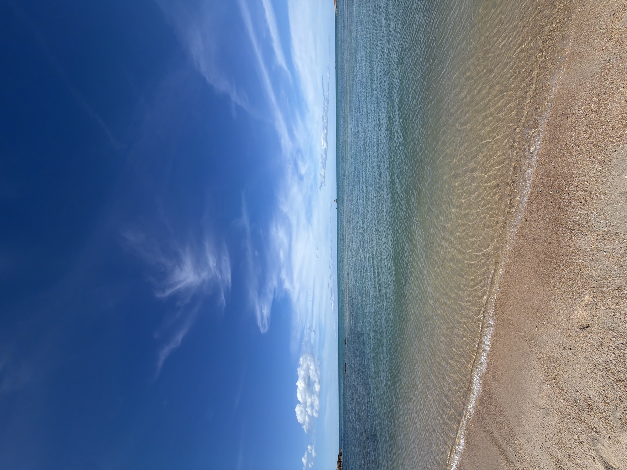This is a panoramic, top-down view of a beach scene in Baan Plai Laem, Thailand. The image is split into two distinct halves: the left side displays a vibrant blue sky dotted with wispy white clouds, while the right side shows the calm, clear ocean water gently lapping at a sandy shore.
The ocean water is shallow and transparent, allowing the textured sandy seabed to be visible. The sand itself is a mixture of light brown and speckled with tiny shells and pebbles. The gentle waves create a shimmering effect on the water's surface, highlighting the sunlight.
In the distance, a faint horizon line separates the sea and sky, with the subtle outline of land or distant structures visible. A few dark, indistinct shapes in the water might be small boats or swimmers, though they are too far away to be clearly identified.
The sky is bright and expansive, suggesting a clear day with pleasant weather, likely mid-day given the brightness. The clouds are delicate and feathery, adding texture to the deep blue canvas. The overall mood is serene and peaceful, evoking a sense of a calm day at the beach. There is no visible text in the image.