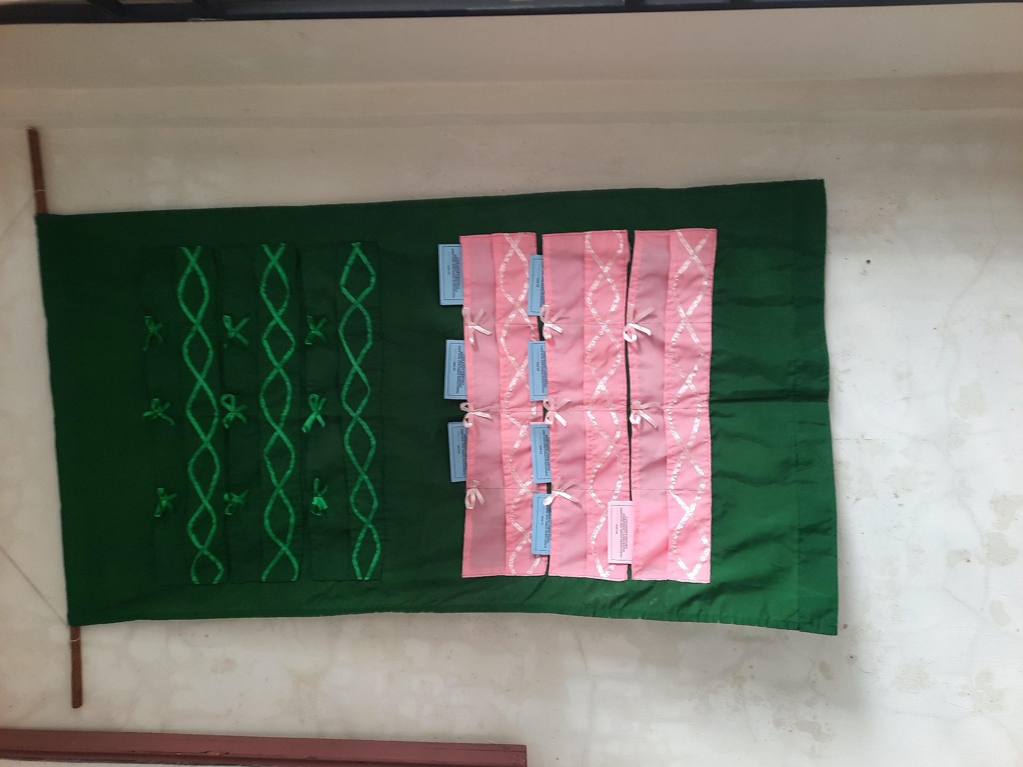 A green fabric wall hanging, decorated with green ribbon in a pattern of intertwined loops and small bows, is displayed on a textured, off-white wall. The lower portion of the hanging features rows of pink fabric pockets, each adorned with a small white bow. Several small, light blue cards with text are inserted into the top pockets. The wall hanging is suspended from a wooden rod. The scene is indoors, suggested by the wall and the presence of what appears to be a window frame on the right edge of the image. The specific location in Lhokseumawe, Indonesia cannot be determined from the visual information provided.
