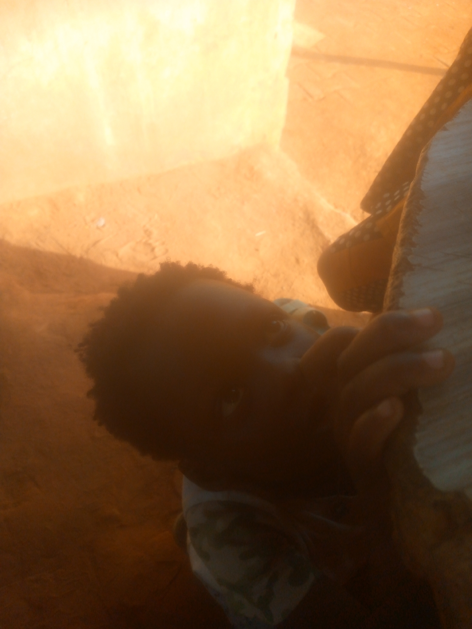 A person with dark skin and curly hair is holding onto a white and brown patterned object. The person's face is partially obscured, but their eyes are visible. The background appears to be an outdoor setting with a wall or structure on the left side. The color palette is dominated by shades of brown, orange, and beige, suggesting a warm or sunny environment. The location is unknown.