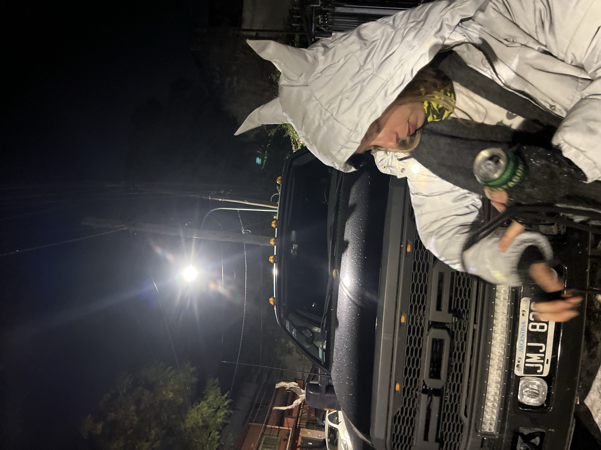 At night in Olivos, Argentina, a person wearing a white, pointed-hooded jacket is seen standing near a black Ford pickup truck. The truck has an Argentine license plate reading "JMJ 82". The person holds a green and black can. A street light is visible in the background.