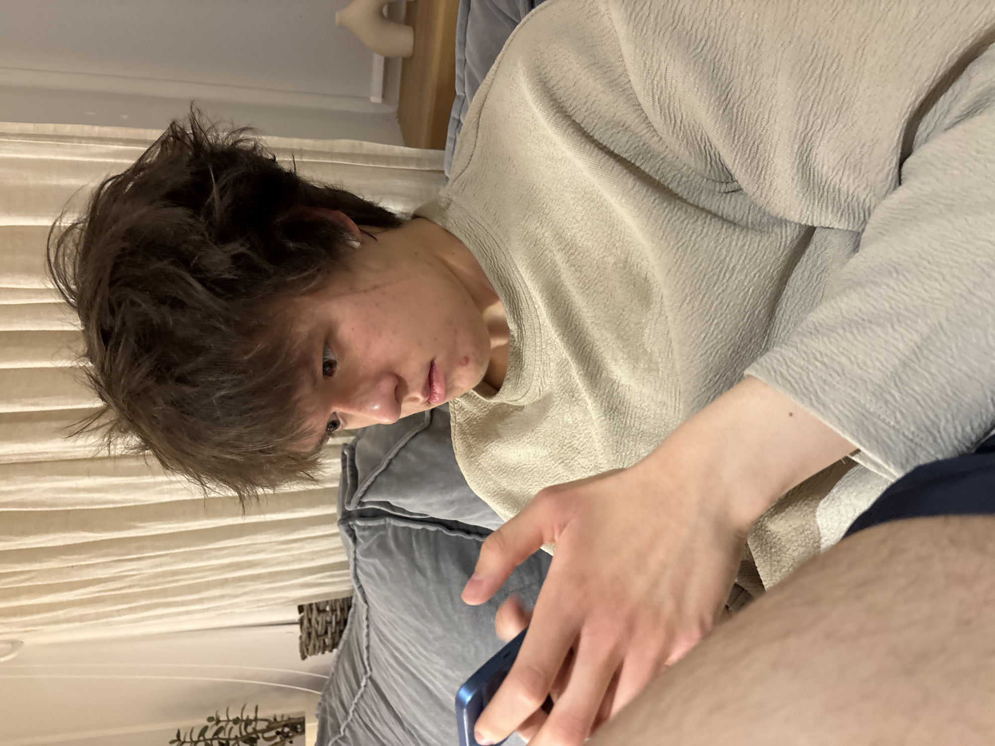 A person with dark brown hair, fair skin, and wearing a beige textured shirt is sitting on a gray cushioned surface. They are looking down at a blue mobile phone in their hand. In the background, there are white horizontal blinds and a wooden shelf with a white object on it. The location is a room, potentially a residential space, possibly in Chaikovsky, Russia.