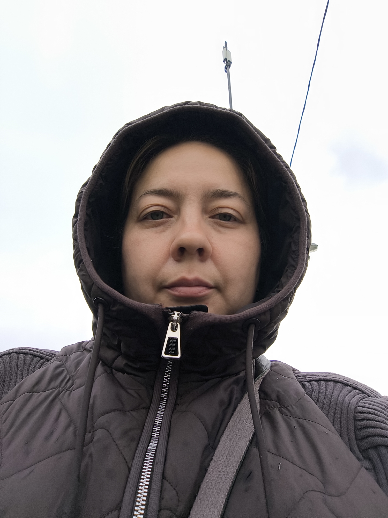 A medium close-up shot of a woman wearing a dark, hooded jacket. The zipper of the jacket is visible. The background is an overcast sky, and there are indistinct structures, possibly a cell tower or utility pole, visible in the distance. The location is Unknown, Unknown.