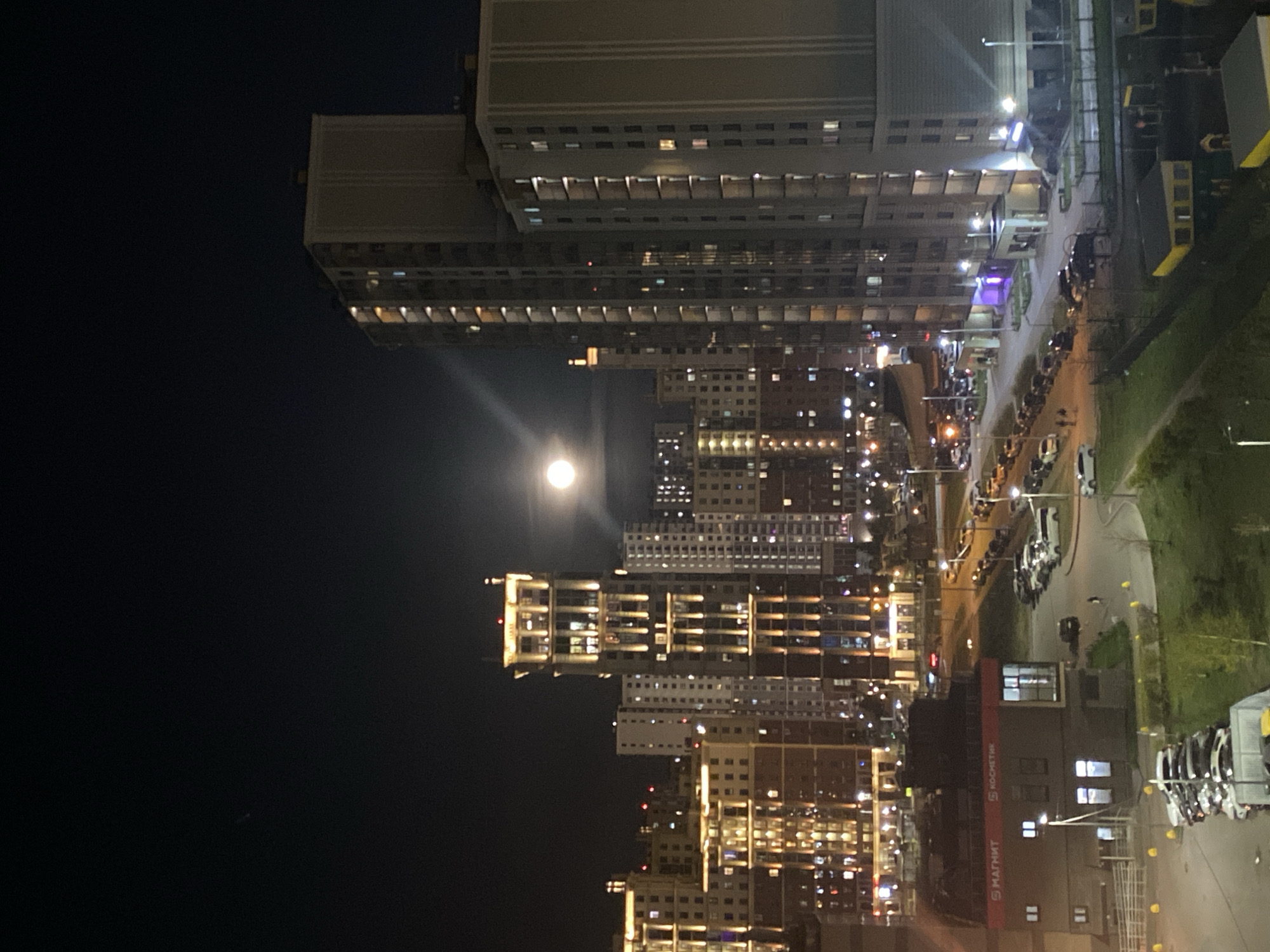 A night view of the city of Saint Petersburg, Russia, shows several high-rise buildings, many with illuminated windows, stretching across the frame. A bright moon is visible in the dark sky. A road with parked cars is visible to the right of the buildings. The words "Kochkethoe" and "Matht" are visible on the side of one building. There are no people visible in the image.