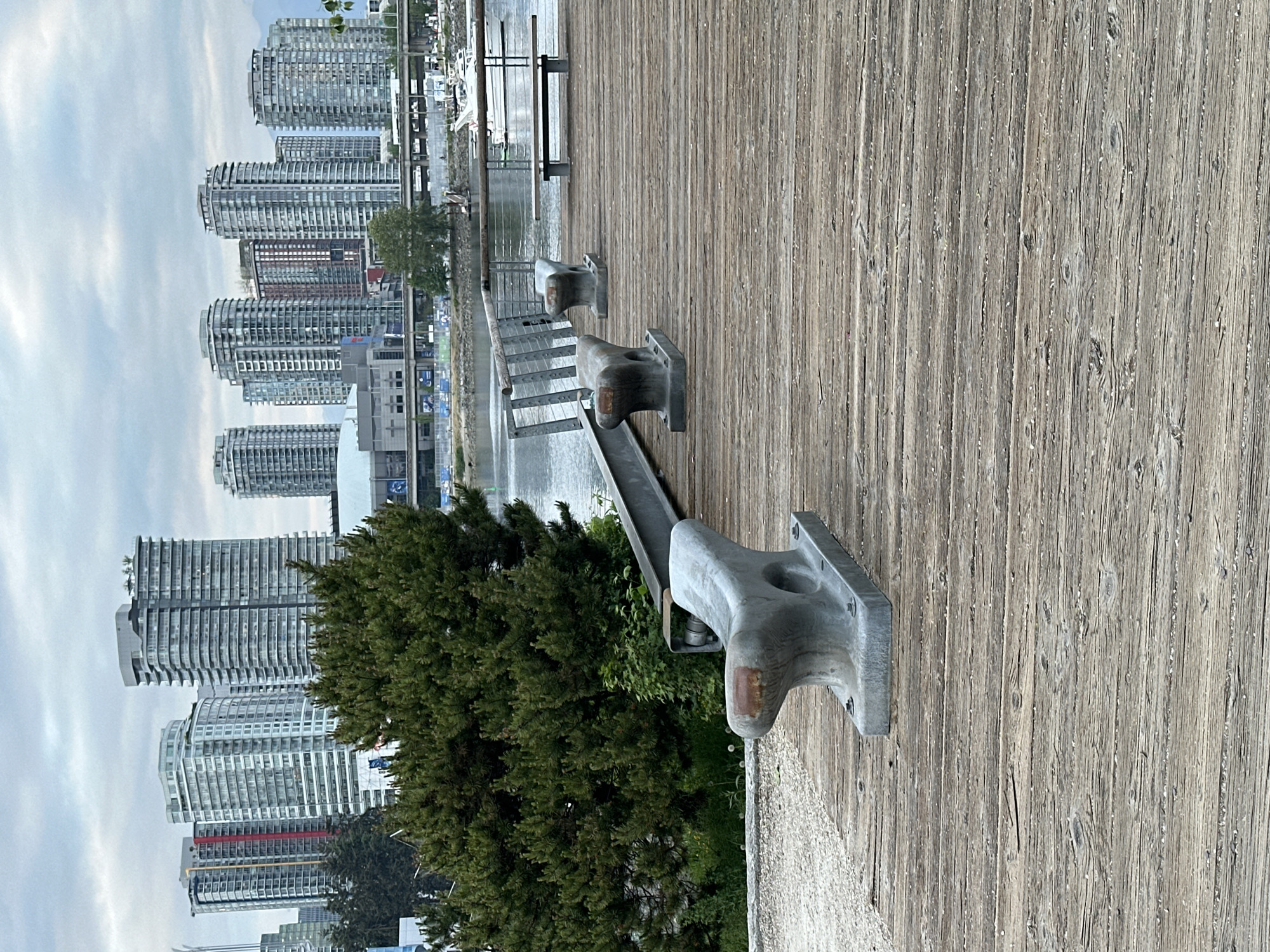 The image depicts a waterfront scene in Vancouver, Canada, featuring a wooden boardwalk with metal mooring bollards. Adjacent to the boardwalk is a metal railing overlooking the water. In the background, there is a cluster of tall, modern apartment buildings that are typical of Vancouver's skyline. A small tree or bush is visible near the mooring bollards, adding some greenery to the scene. The sky is overcast.