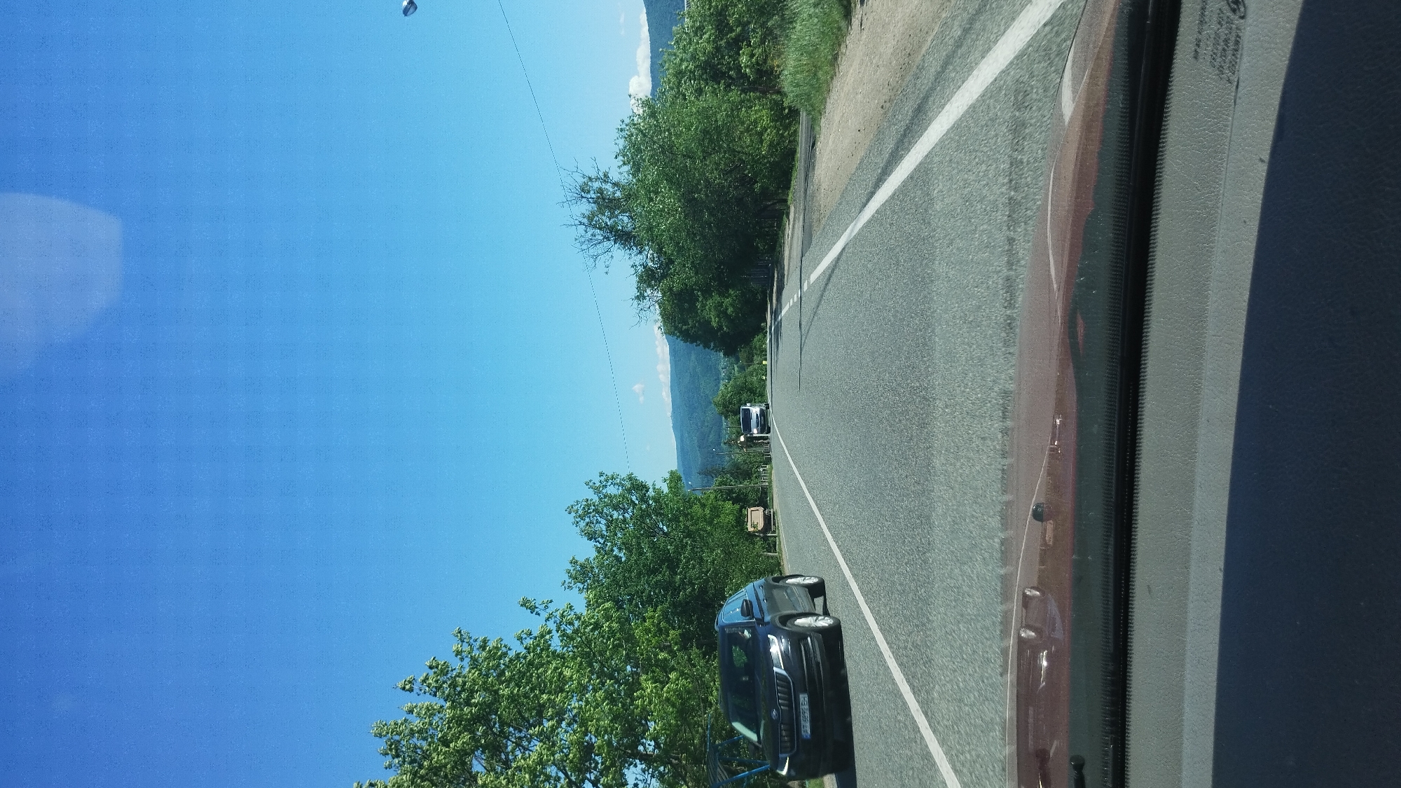 The image shows a road in Deliatyn, Ukraine, from the perspective of a vehicle. A black car is driving on the road, and another vehicle is visible further ahead. Trees line the side of the road. The sky is blue. Mountains are visible in the distance.