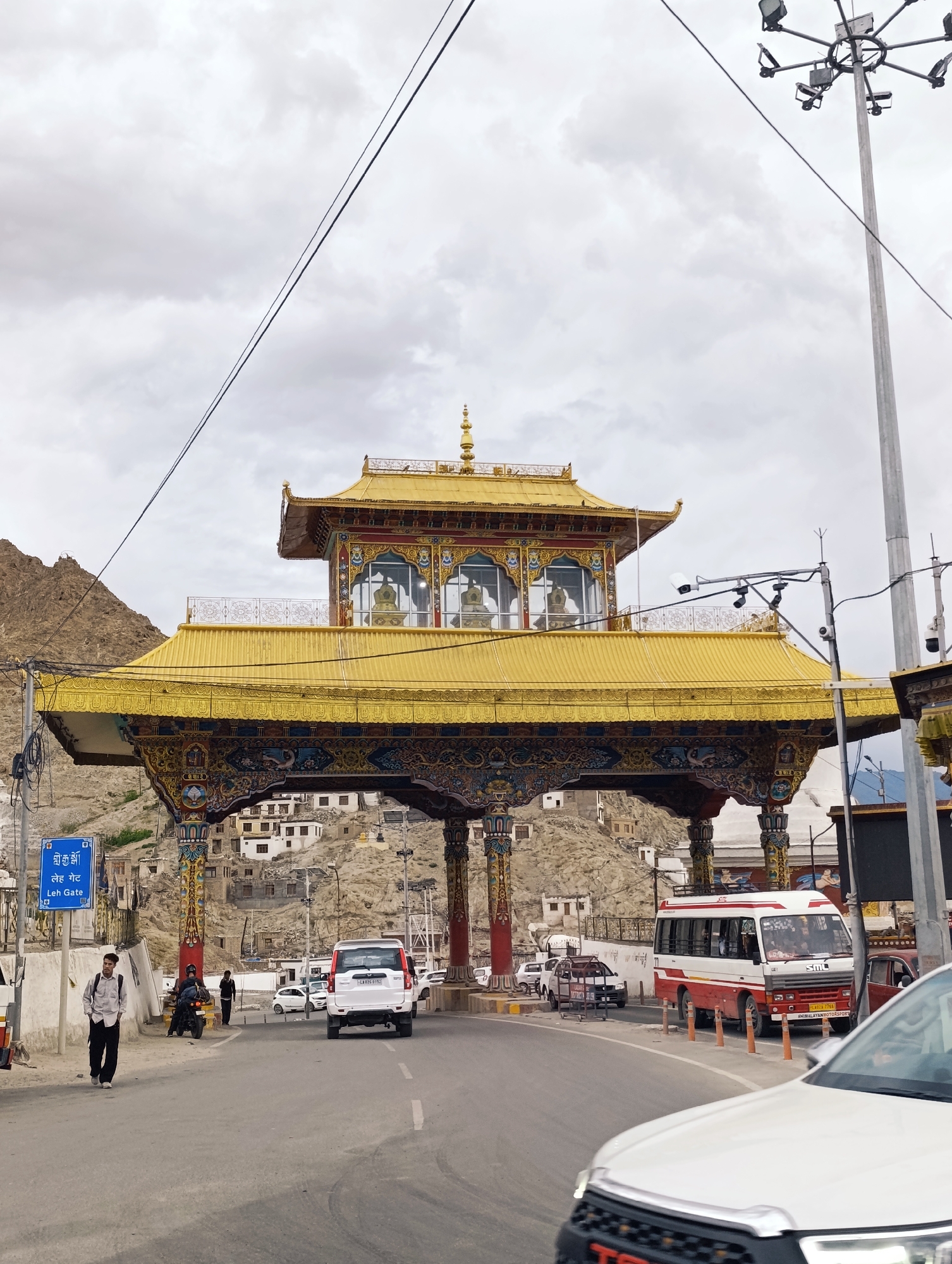 A white car drives on a road in front of a large, ornate golden gate in Leh, India. The gate is topped with a tiered, golden roof and has colorful, detailed carvings on its supports. Behind the gate, a hillside is dotted with white buildings. The sky is overcast with gray clouds. A red and white bus is parked to the right of the gate. People are seen walking and riding a motorcycle on the road. A blue sign with text in Tibetan and English, reading "Leh Gate," is visible to the left of the road. The scene appears to be a point of entry or a significant landmark in Leh.