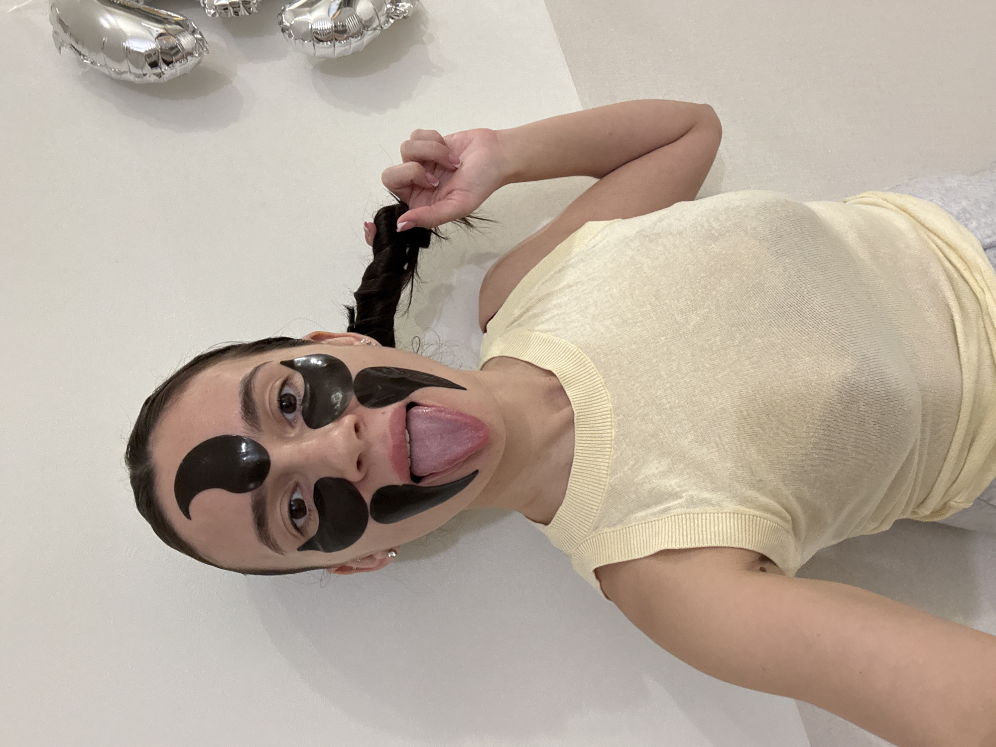 A woman is lying down, wearing black eye masks and sticking out her tongue. She is wearing a light yellow shirt and holding a strand of dark hair in her hand. There are silver balloons visible in the top left corner of the frame. The background is a white wall. The location is Chaikovsky, Russia.