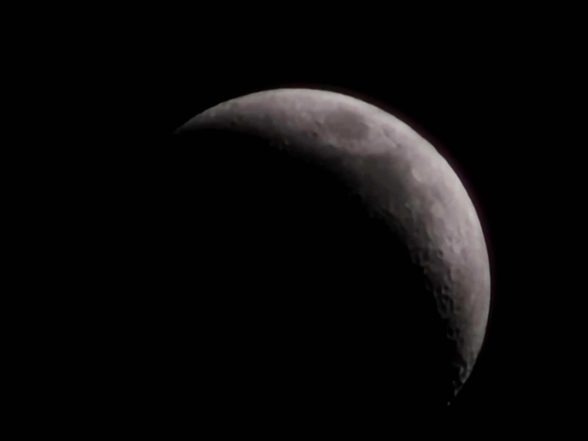 The image displays a close-up view of the Moon in a distinct crescent phase, set against a deep, dark, starless night sky. The illuminated portion of the Moon, appearing on the right side of the frame, reveals a detailed grey and white surface textured with visible craters and lunar features, particularly along the terminator line. The curvature of the crescent is sharp and well-defined. A subtle, faint reddish or purplish halo is visible around the luminous edge of the Moon. The remaining, unlit portion of the lunar disk blends into the absolute blackness of the surrounding space. There are no other celestial bodies visible. The scene depicts an astronomical observation during nighttime, implying clear weather conditions. No activity, emotions, or visible text are present in the image. The location context provided is Esch-sur-Alzette, Luxembourg.