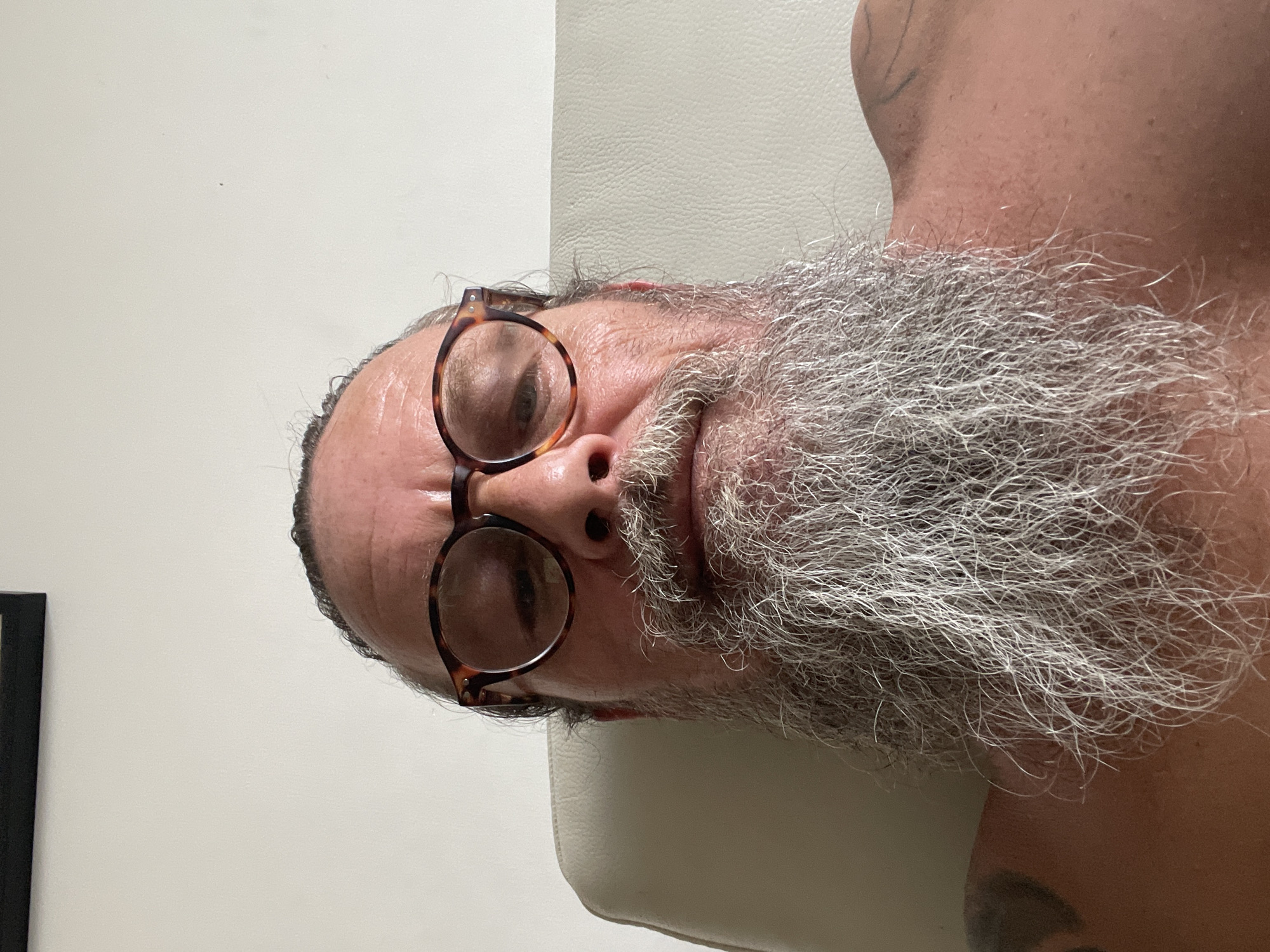 The image is a close-up, low-angle selfie of a person with a long, gray beard and glasses. The person appears to be sitting in a light-colored chair, possibly made of leather, against a plain white wall. A dark frame is visible at the top of the image, suggesting a picture or window. There are tattoos on the person's shoulder and upper back. The overall setting is indoors and appears to be a home or a similarly private space. The image was taken in Netanya, Israel.