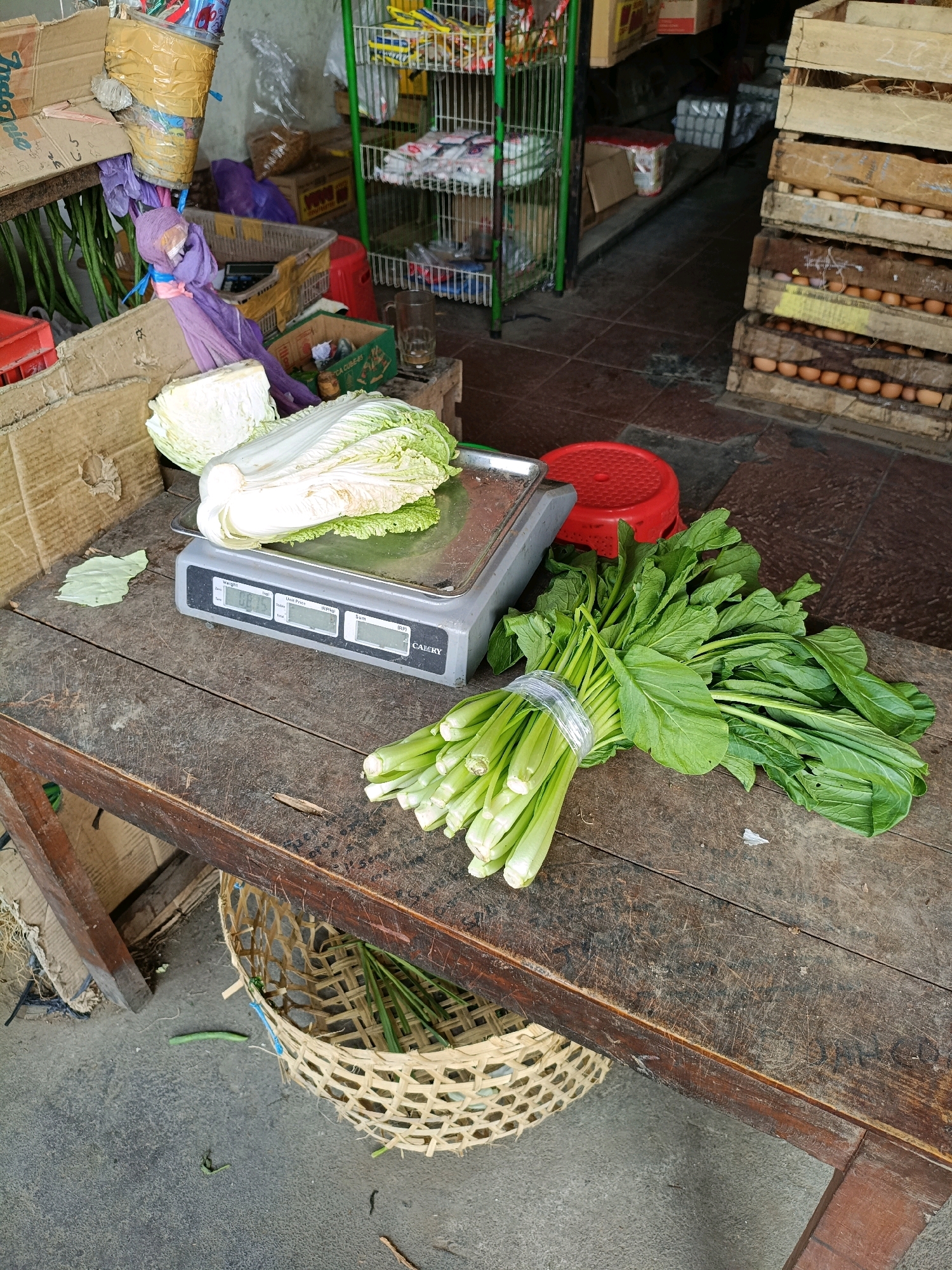 The image depicts a scene at a street market stall in Maguwoharjo, Indonesia. In the foreground, a weathered wooden table holds a digital scale with a head of napa cabbage placed on it. To the right of the scale, a bunch of leafy green vegetables, possibly mustard greens or bok choy, is tied with clear plastic wrap. Behind the table, a woven bamboo basket sits on the ground, containing some long green stalks, possibly more vegetables. In the background, various items are visible, including cardboard boxes, a red plastic stool, shelves stocked with packaged goods, and wooden crates filled with eggs. The overall environment suggests an active but somewhat cluttered market setting. There are no people visible in the image.