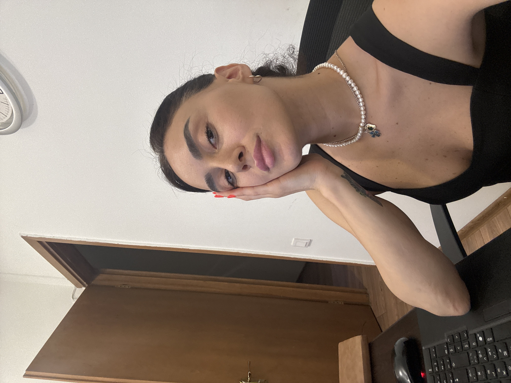 The image is a medium shot of a young woman with dark hair pulled back, sitting at a desk in an office. She is looking towards the camera, with her left hand resting on her cheek. She is wearing a black top and a pearl necklace with a blue and silver pendant. A computer keyboard and mouse are visible on the desk in front of her. To her left, a wooden door is ajar, revealing a dark interior. The wall behind her is white, with a clock mounted high up. The scene appears to be an indoor office setting. There is no information in the image to suggest it is located in Tbilisi, Georgia.