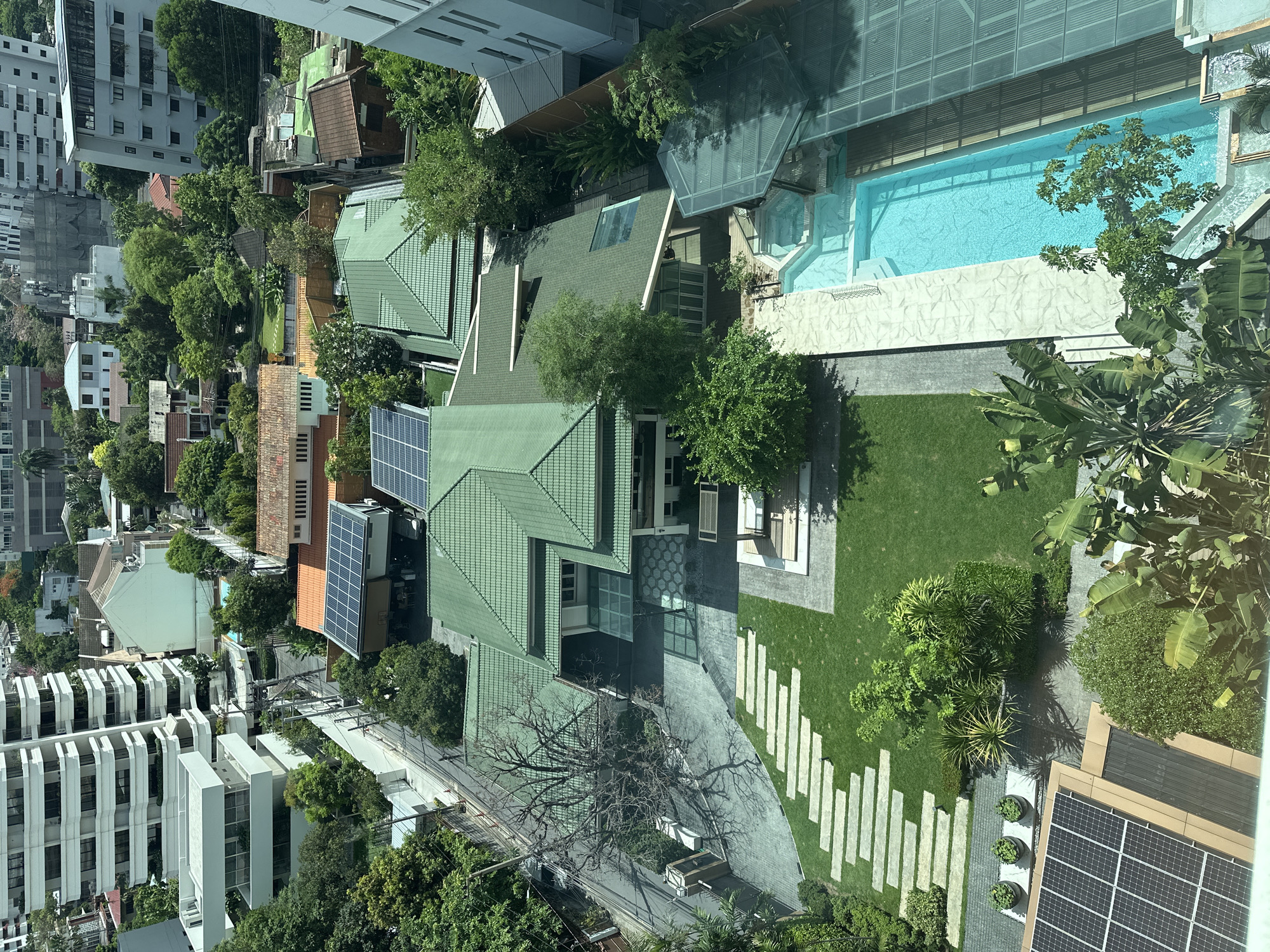 An aerial view captures a residential area within the city of Bangkok, Thailand. The architecture includes a combination of high-rise buildings and individual houses, most of which feature green rooftops. Some houses have solar panels installed on their roofs. A large swimming pool is visible in the upper right quadrant of the image. Trees and greenery are abundant, interspersing the buildings and adding to the urban landscape. A unique stone pathway of individual stone is visible leading to the house.