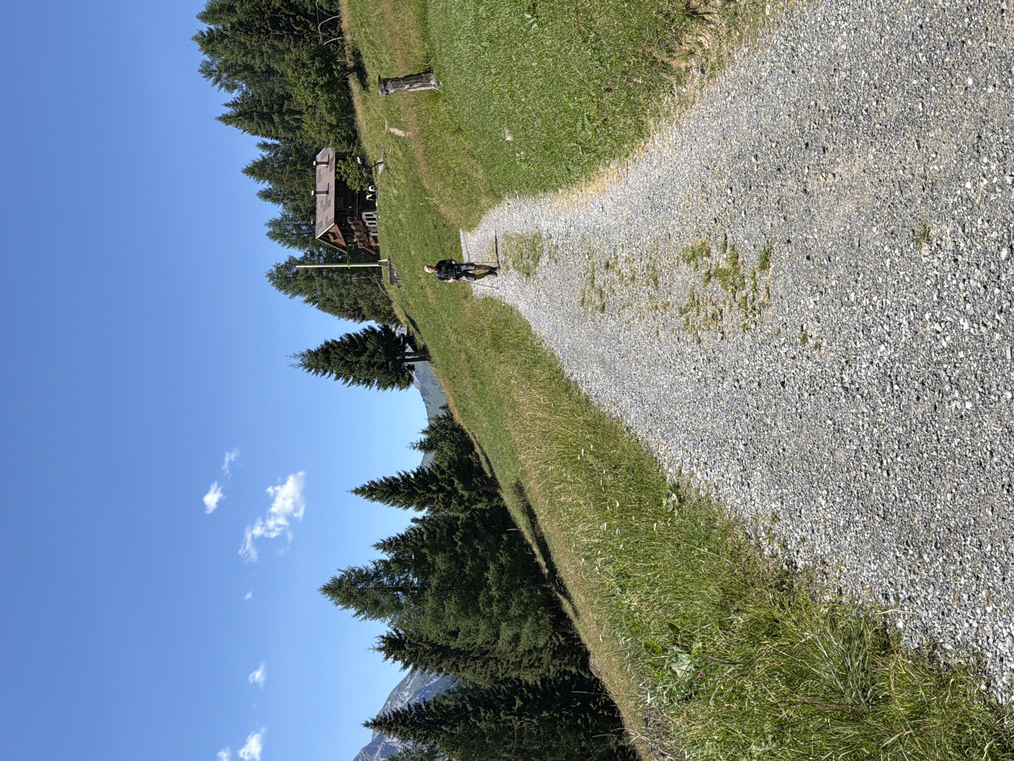 A lone hiker with trekking poles walks up a gravel path on a sunny day. The hiker is wearing a dark t-shirt, grey shorts, and a hat. The path curves uphill, with lush green grass on either side. In the background, a traditional wooden house with a red roof is nestled on a hillside surrounded by tall evergreen trees. Beyond the trees, a bright blue sky with a few scattered white clouds is visible. The image appears to depict a rural or mountainous setting in the Italian Alps, possibly in the region of Lateis / Latais.