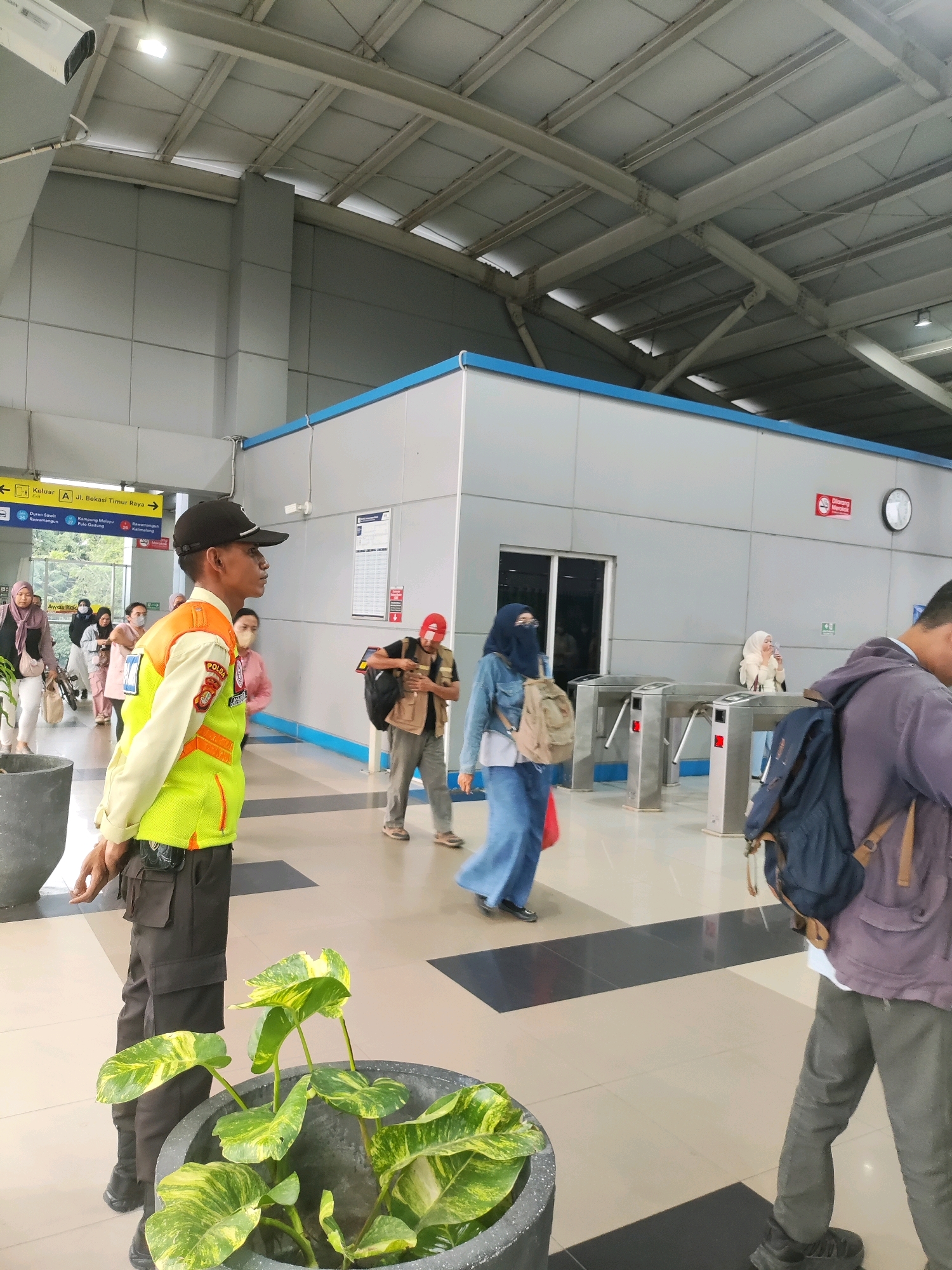 The image depicts a scene at a train station in East Jakarta, Indonesia. In the foreground, a security guard wearing a black cap, a light-colored uniform shirt with a bright green and orange vest, and dark pants stands facing to the right. He has a black pouch attached to his belt. To the left of the guard, a large potted plant with variegated green leaves is visible.
In the mid-ground, several individuals are walking through the station. One woman, wearing a dark hijab and a denim jacket, is walking towards the viewer with a backpack. Another person in a red cap and a beige vest is adjusting a backpack. A woman in a light blue headscarf and a long blue dress is seen walking away from the viewer towards a set of ticket barriers. Behind her, two women in hijabs are walking together.
The background features a modern station interior with a silver-colored wall and a blue trim along the top. Ticket barriers are visible, and a clock is mounted on the wall. Overhead, the station's roof structure with exposed beams and lighting is apparent. Signage indicating "Keluar" (Exit) and directions to "Jl. Bekasi Timur Raya" and "Kampung Melayu Pulo Gadung" are also present. The flooring is tiled with a pattern of beige and dark gray squares.