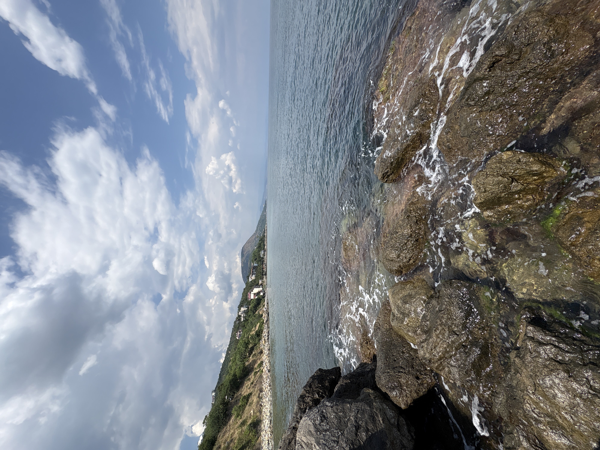 The image depicts a scenic coastal view with large rocks in the foreground, lapped by calm, blue water. Beyond the water, a tree-covered hillside slopes up, dotted with white buildings. The sky is a mix of blue and white clouds. The scene is located in Bondarenkovo, Ukraine.