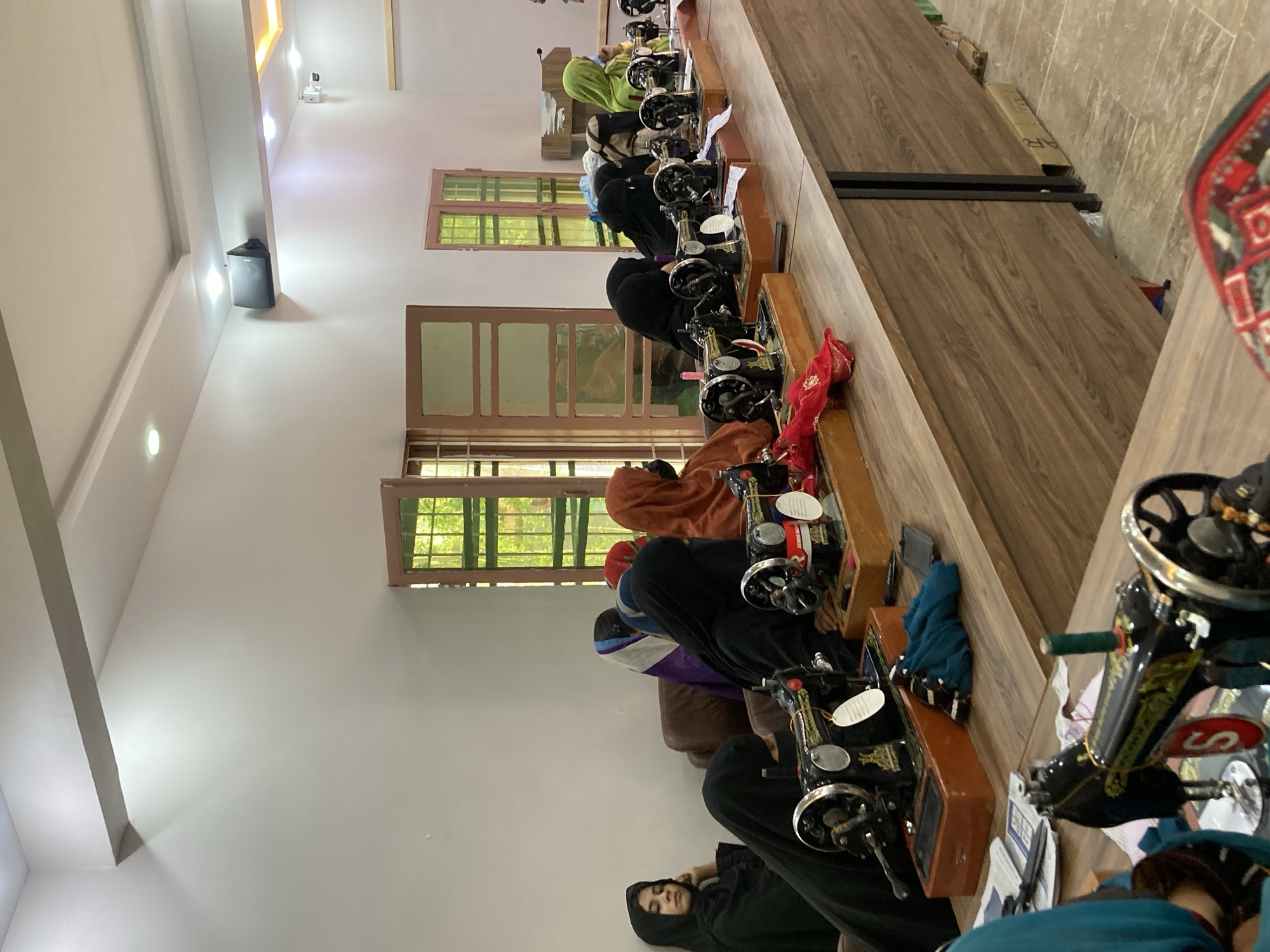 Inside a brightly lit room in Rāwāni, Pakistan, a row of women, some wearing headscarves, sit at individual sewing machines positioned along a long wooden table. Each woman is focused on their work. Windows line the wall behind them, allowing natural light to flood the room. A speaker is mounted high on the wall. The sewing machines are vintage, likely manually operated. The scene suggests a vocational training center or small-scale garment workshop.
