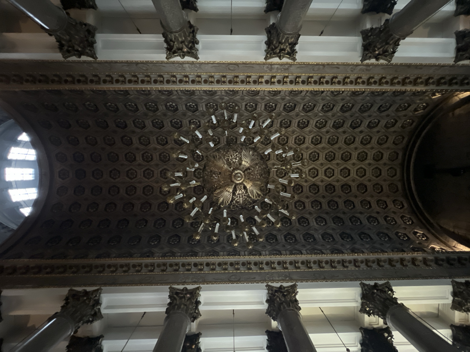 The image depicts an ornate ceiling with a large chandelier in the center, likely taken inside a historic building. The ceiling features a pattern of hexagonal shapes, each containing a floral design. The chandelier is multi-tiered with numerous lights that resemble candles. The ceiling is framed by decorative molding. The building also features tall columns with ornate capitals. A window is visible on the left and an archway is visible on the right. Based on the architectural style and details, the image is likely of the interior of Saint Isaac's Cathedral in Saint Petersburg, Russia.