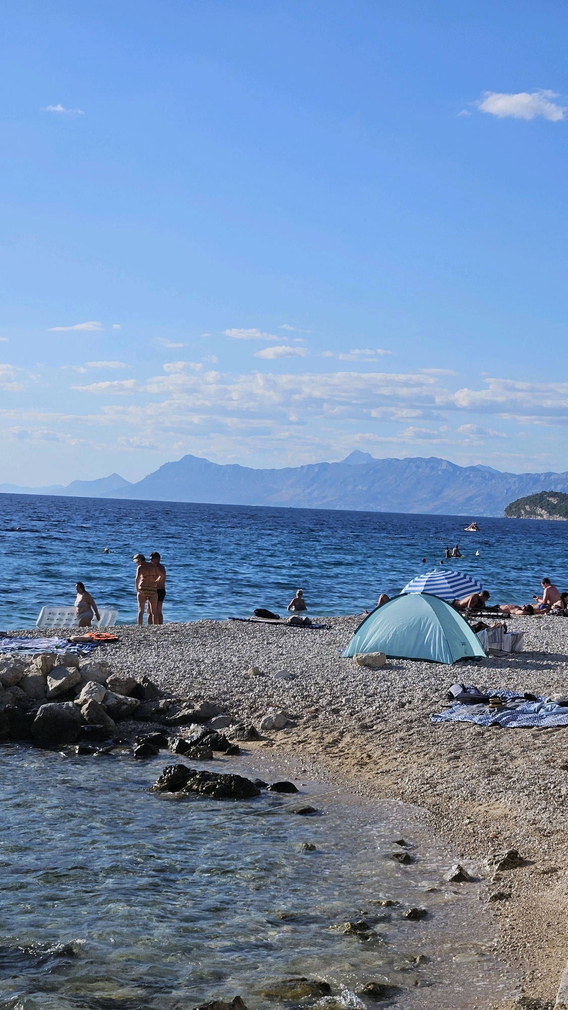 The image depicts a sunny day at a pebble beach in Općina Tučepi, Croatia. The foreground shows clear, shallow water lapping at the shore, with submerged rocks visible. The beach itself is covered in a mix of pebbles and sand. Several people are present: two individuals stand together in the water near the left side of the frame, one in a patterned bikini and the other in a dark swimsuit. Further back, near the water's edge, another person in a bikini is walking with an inflatable mattress. In the mid-ground, a light blue tent is set up on the beach, alongside a blue and white striped umbrella. Several individuals are relaxing on towels or loungers near the tent. The background features the vast, blue Adriatic Sea, with distant mountains rising majestically on the horizon, their peaks partially shrouded in haze. A few boats are visible on the water. The sky is a vibrant blue with scattered white clouds.