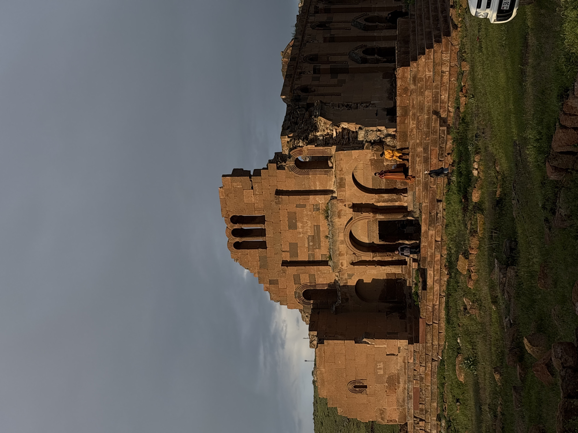The image shows the ruins of a stone building, likely a church or similar structure, located in what appears to be a grassy field. Three people are standing or walking on the stone structure. One is wearing orange or yellow clothing and another wearing red clothing. The sky is overcast with a light grey color. A white vehicle is partially visible on the upper right. The location is possibly in the city of Anipemza, Armenia.