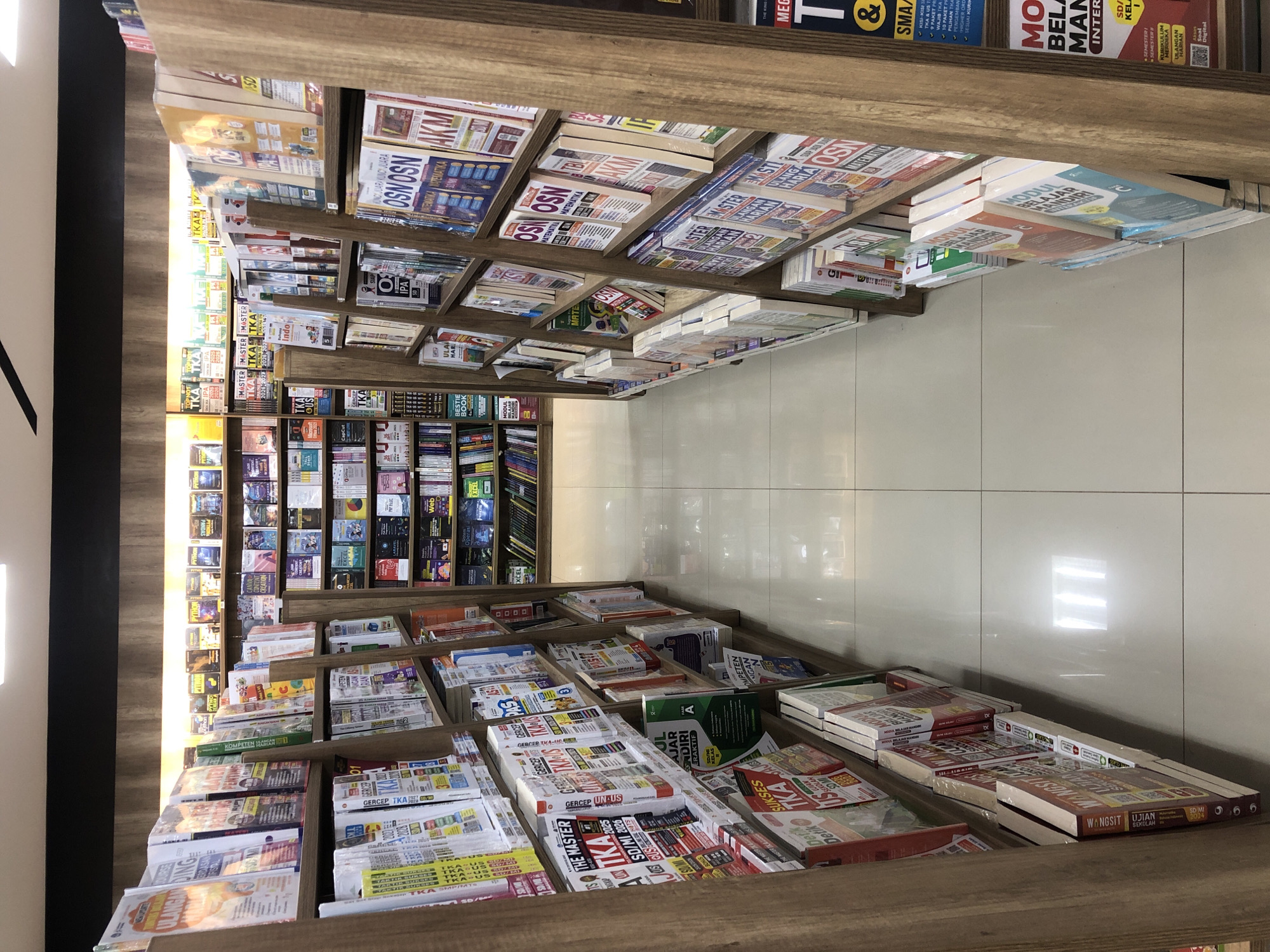 This image shows a narrow aisle between two tall bookshelves in a bookstore, likely in Banda Aceh, Indonesia, given the content of the books which appear to be in Bahasa Indonesia and cater to educational subjects. The bookshelves are constructed of light brown wood. The books are neatly arranged on the shelves, with their spines facing outwards. The majority of the books are textbooks or study guides, with titles suggesting subjects like mathematics, science, and general education. Some books have bright, colorful covers, while others are more subdued. The aisle floor is covered with large, light-colored square tiles. The lighting is bright, coming from overhead fluorescent lights, creating reflections on the floor. There are no people visible in the image. The scene is clean and orderly, typical of a retail environment focused on educational materials.