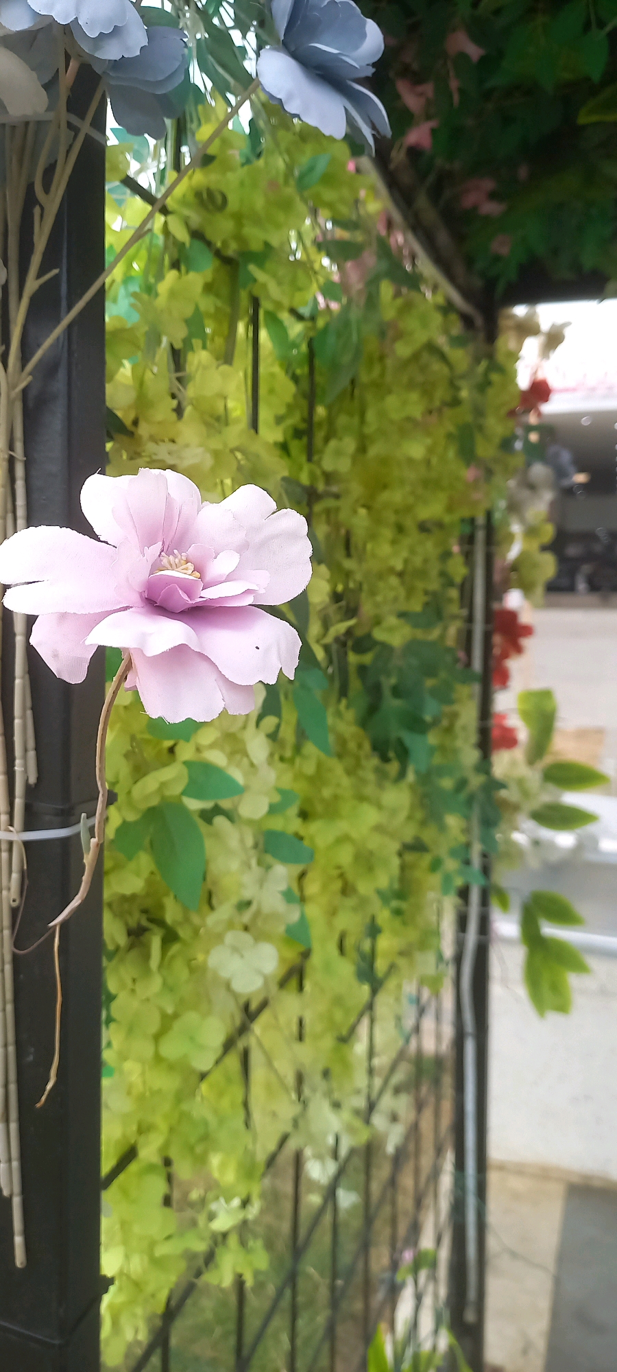 A close-up, eye-level shot features a single, delicate pink artificial flower prominently in the foreground, its petals slightly unfurled. The flower is attached to a thin stem that extends downwards and slightly to the left. To the left of the flower, a dark, vertical pole is visible, with thin, light-colored, hollow tubes bundled together and attached to it with a white zip tie. This pole and the flower appear to be part of an artificial floral arrangement, possibly decorative.
Behind the flower and pole, a dense, vibrant green foliage, likely artificial, fills the mid-ground. This foliage is characterized by numerous small, rounded leaves and some scattered lighter green clusters that could be smaller blooms or buds. Interspersed within the green are a few small, artificial red flowers. The foliage is draped over and around a black metal fence with a diamond-shaped mesh pattern.
In the background, the scene opens up to an outdoor environment. A white-railed structure runs horizontally across the right side of the frame. Beyond the fence and foliage, the blurry shapes of buildings and vehicles suggest an urban or commercial setting. The overall lighting is bright, indicating daylight. The composition is slightly tilted, with the flower on the left providing a focal point against the more generalized, blurred background. There are no visible people or animals in the frame. The image appears to be taken in a public space, possibly a park, garden, or the exterior of a business.