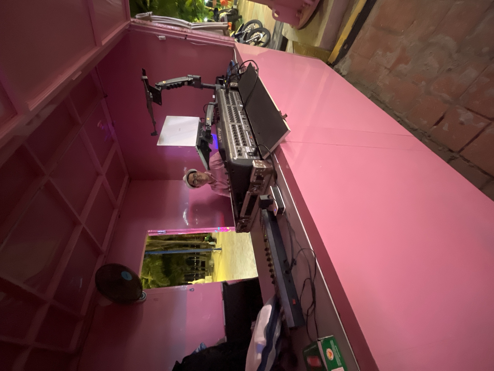 A person is seated behind a pink counter, wearing a pink shirt and a white hat, with glasses. They are looking towards the camera. In front of them is a large audio mixing board with various knobs and connections, and a laptop is mounted on a stand above it. To the left of the mixing board is a black cable connected to a power strip, and further left is a small green box. Behind the person, an opening reveals an outdoor scene with palm trees and a beach area, suggesting a location in a coastal city like Đà Nẵng, Vietnam. The interior of the counter area is painted pink, and a black fan is mounted on the ceiling. To the right of the counter, two motorcycles are parked. The flooring appears to be made of brick. The overall scene suggests a DJ or sound technician operating equipment in an open-air venue.