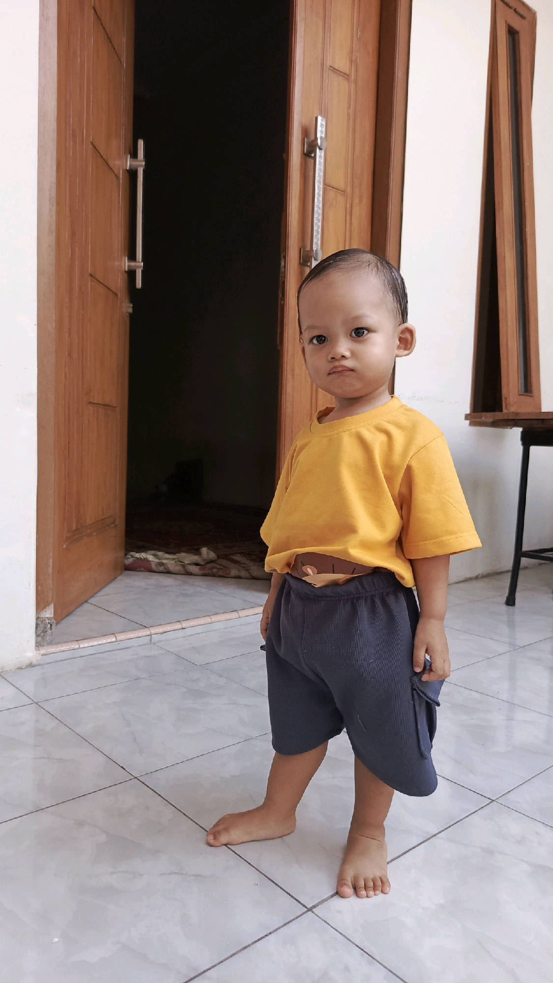 A young child stands barefoot on a tiled floor in front of an open wooden door. The child is wearing a bright orange t-shirt and dark gray shorts. The door, made of wood with vertical paneling, has two silver handles and is slightly ajar, revealing a dark interior and a patterned rug on the floor. To the right of the child, a portion of a brown wooden structure and a black metal stool are visible. The environment appears to be an outdoor or semi-outdoor space, possibly a veranda or entrance area of a building in Pekauman Kulon, Indonesia.