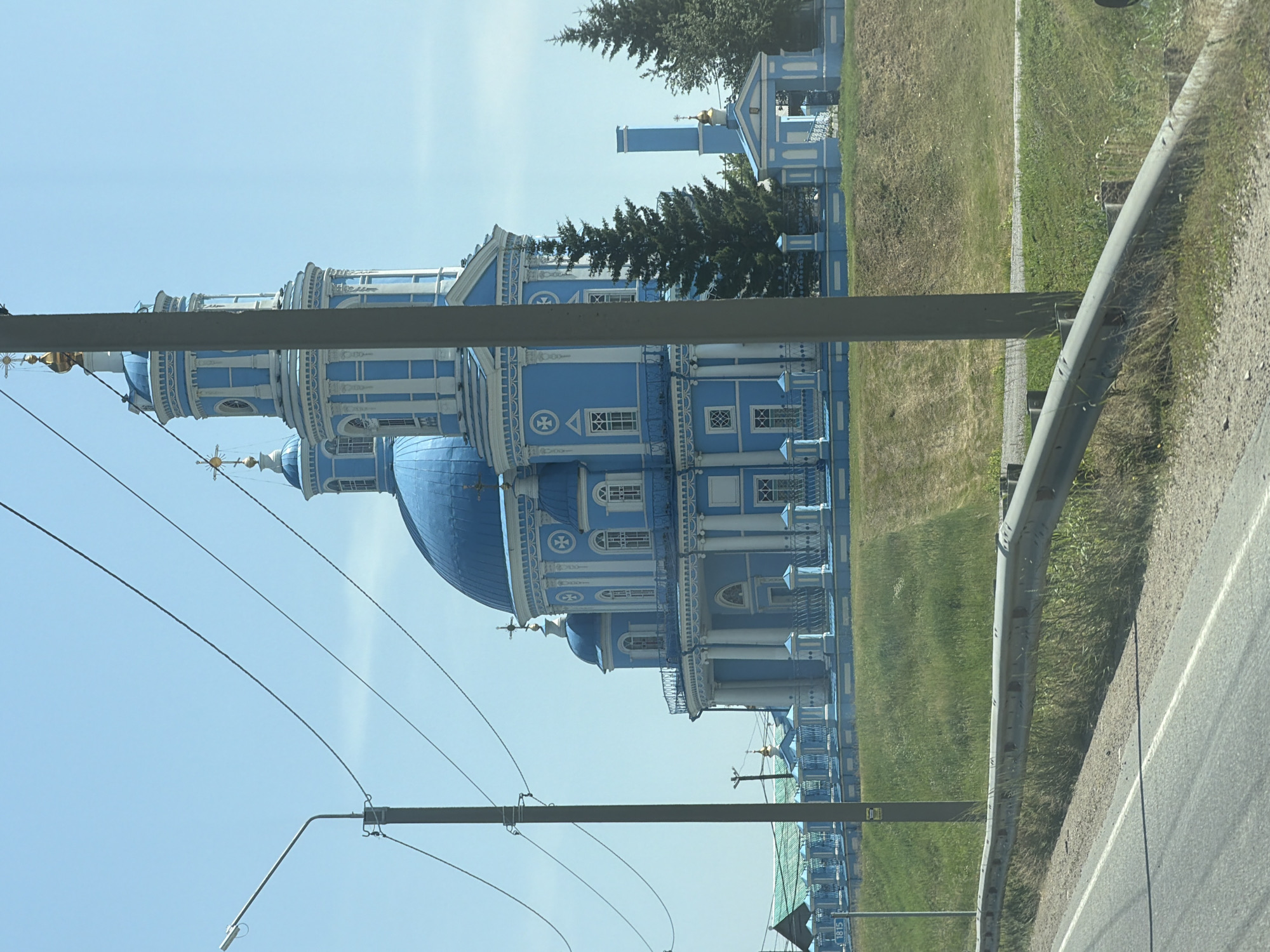 The image captures a grand blue and white church with multiple domes and intricate architectural details, possibly in Telma, Russia. A tall clock tower with a golden cross at its pinnacle dominates the skyline. The church is surrounded by a light blue fence and manicured green lawns. Tall pine trees are visible to the right of the church, and a bright blue sky serves as the backdrop. In the foreground, a gray guardrail separates a paved road from the grassy embankment leading up to the church. Electrical wires stretch across the frame, supported by utility poles, indicating the presence of infrastructure near the religious site. The scene appears to be captured during daylight on a clear day.