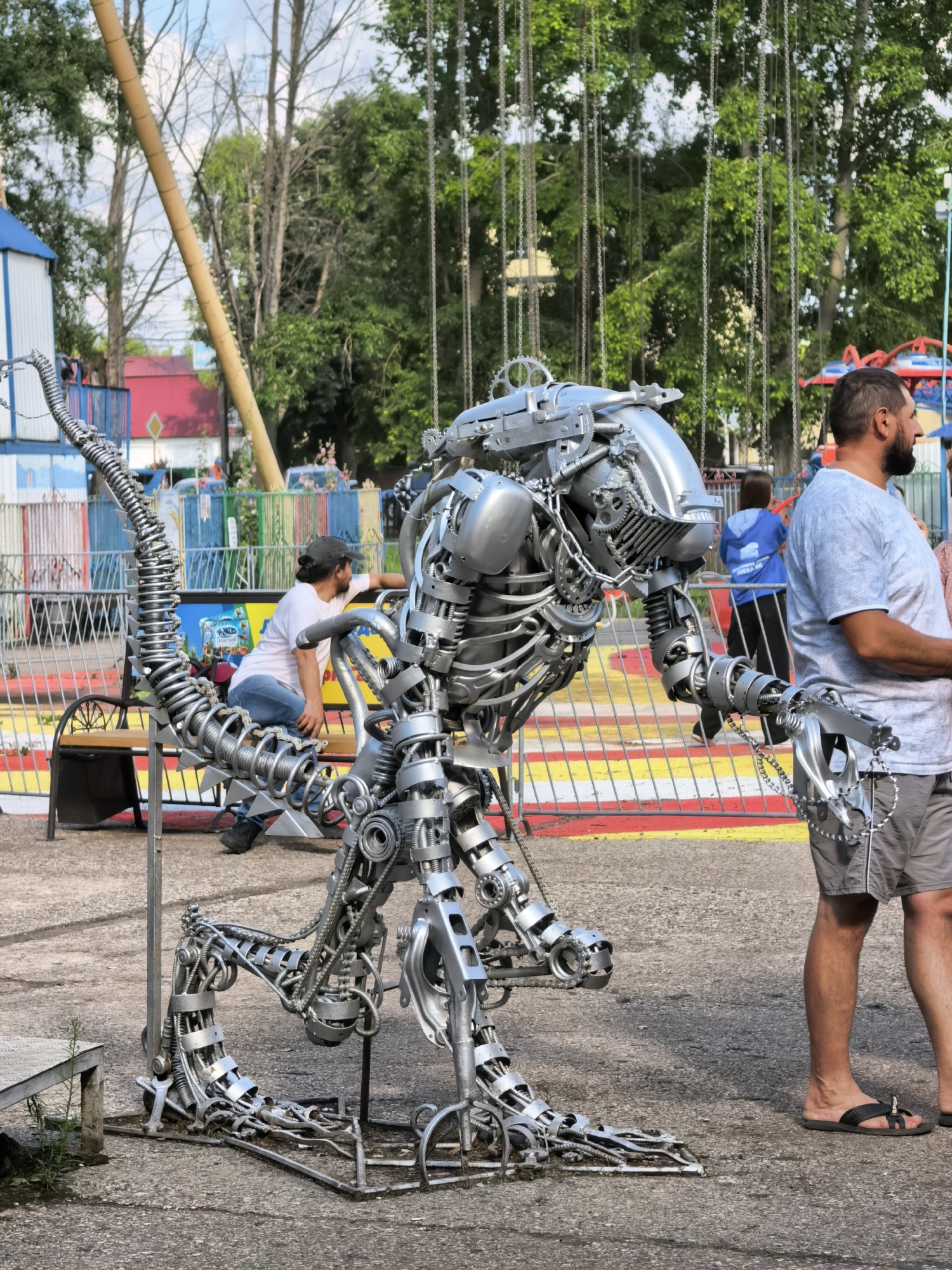 The image depicts an outdoor scene, likely an amusement park or fairground, with a large, intricate metal sculpture of a creature, possibly a dinosaur or robot, in the foreground. The sculpture is made of various metallic components, including gears, chains, and pipes, and is predominantly silver in color. To the right of the sculpture stands a man wearing a light blue t-shirt and grey shorts, with his left arm resting on the sculpture. In the background, a chain-link fence with red and yellow horizontal bars separates the foreground from a seating area and further attractions. Behind the fence, people are visible, including a person in a white t-shirt and jeans crouching on a bench, and others in the distance near amusement rides. Trees and a bright blue sky with some clouds form the backdrop, suggesting a daytime setting. A tall, slender yellow pole is also prominent in the left side of the background. The location is identified as Sterlitamak, Russia.