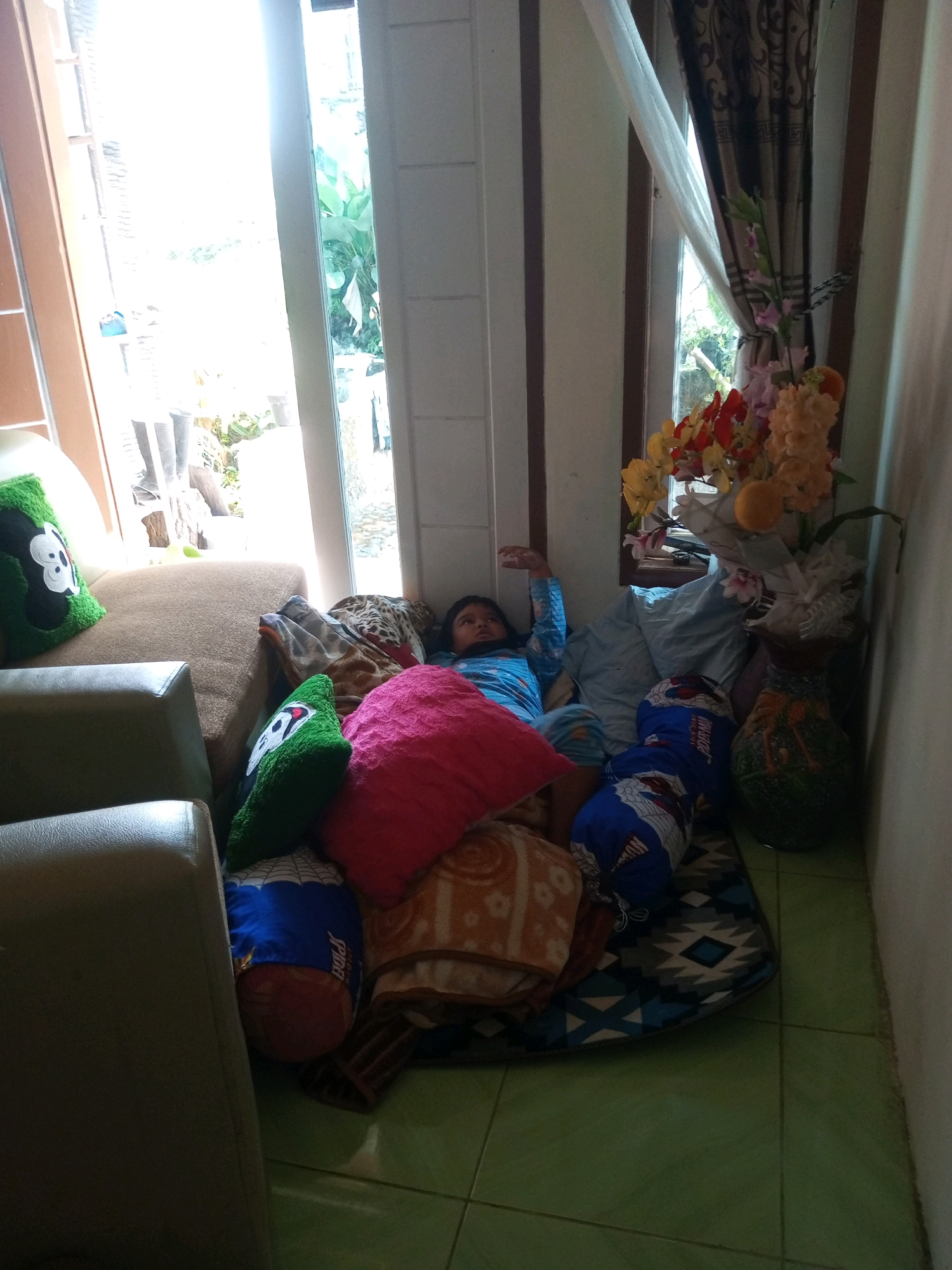 A child is lying on the floor surrounded by pillows and blankets in a room with green tiled floors. The child is wearing a blue long-sleeved shirt and blue pants. To the left of the child, there is a tan couch with a green pillow with a Mickey Mouse design. To the right of the child, there is a vase of artificial flowers. The room appears to be residential. The image is taken indoors.
