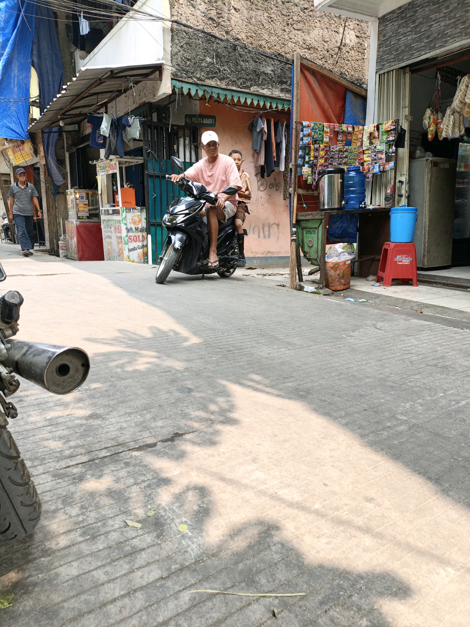 The image depicts a street scene in West Jakarta, Indonesia, likely in a densely populated area. In the foreground, the exhaust pipe of a motorcycle is visible, suggesting the viewpoint is from another motorcycle. In the mid-ground, a man wearing a pink t-shirt and a white cap is riding a black scooter, with another person sitting behind him. They are on a paved street lined with small shops and stalls. To the left, a man in a striped shirt and dark pants is walking away from the camera. The shops display various goods, including clothing and packaged food items. Tarps and awnings cover some of the stalls, and laundry is hung out to dry. The buildings are close together, characteristic of urban residential or commercial areas in Indonesia. The overall atmosphere appears to be that of a busy, everyday street in a local market or neighborhood.