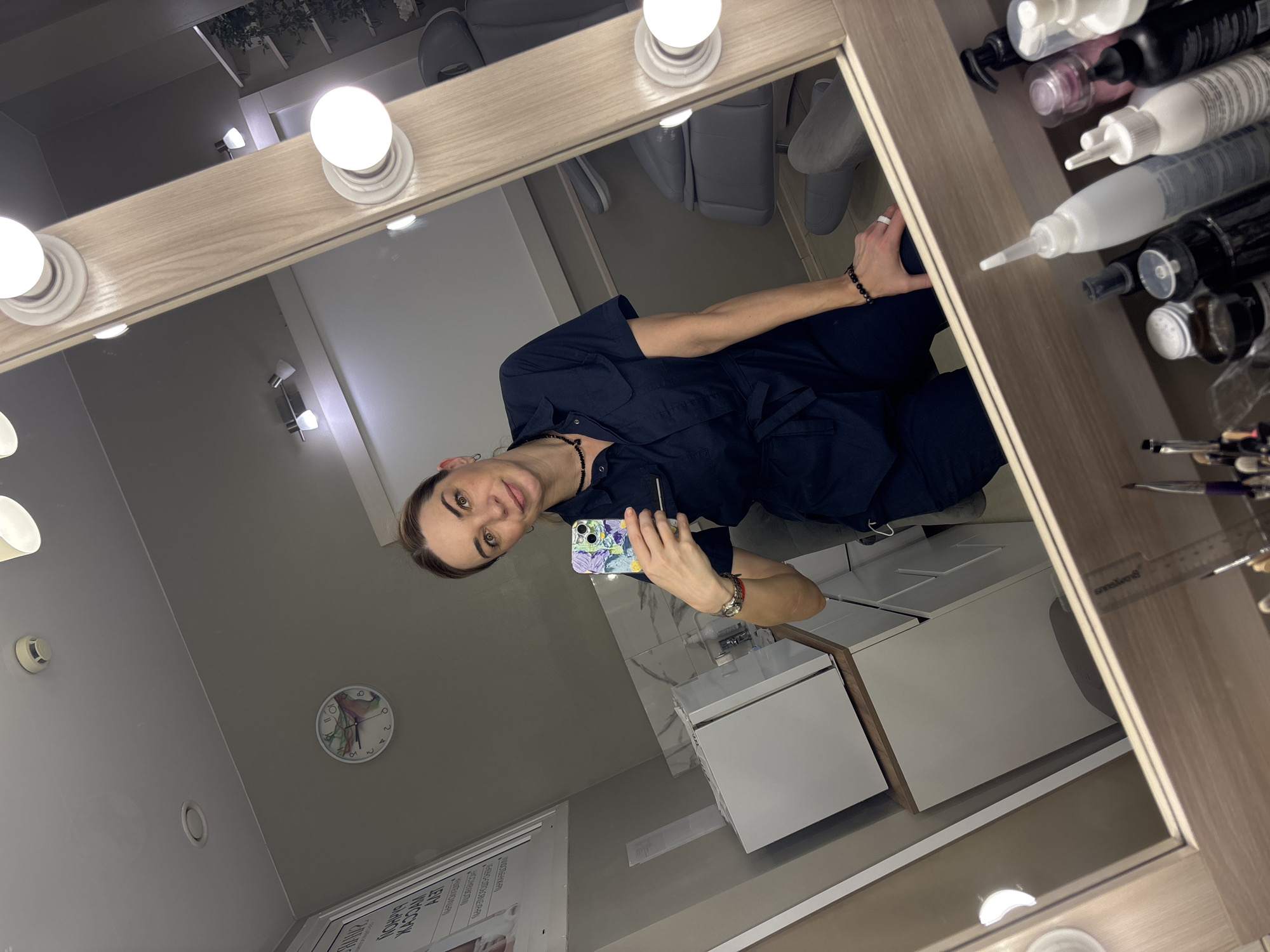 The image depicts a selfie of a person reflected in a makeup mirror. The person is holding a smartphone with a patterned case and wearing a dark blue uniform. The mirror has light bulbs around its perimeter and is positioned in a room that appears to be a beauty salon or makeup studio.
The environment includes:
- Storage units with white doors.
- Shelves holding various bottles and makeup brushes.
- A clock on the wall in the background.
The image was taken in Murino, Russia.