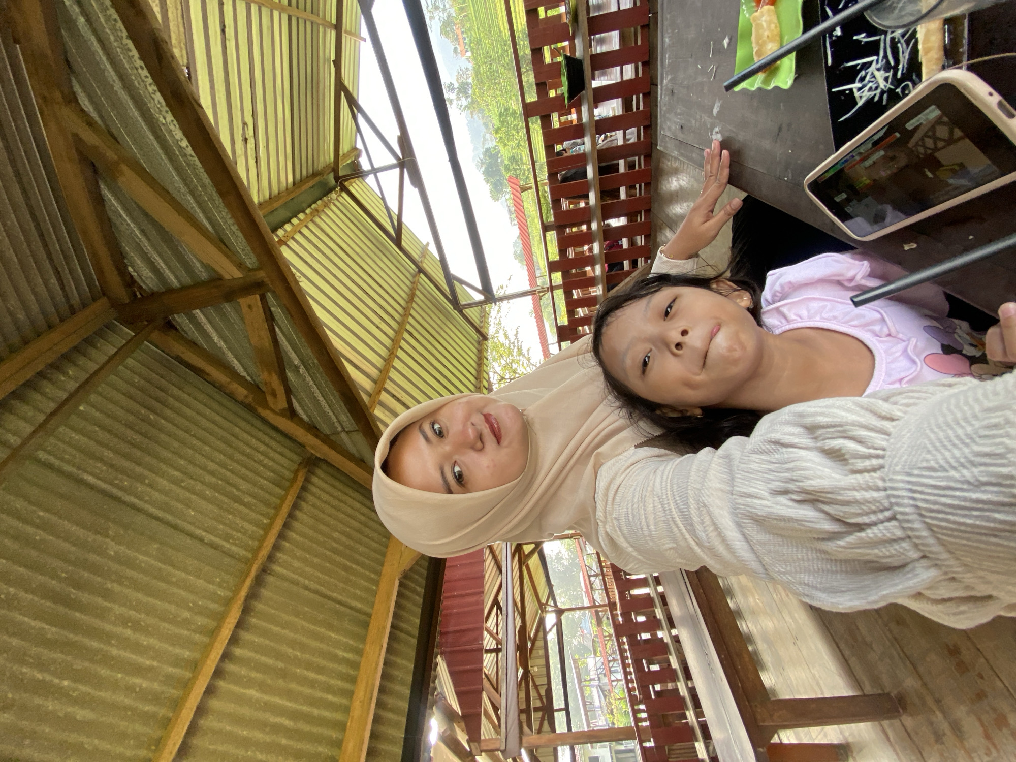 A rotated, candid, outdoor shot captures a woman and a young girl. The woman, positioned on the left, wears a tan-colored hijab. The girl, located on the right, has black hair and wears a pink shirt. They are both holding a black phone. The setting seems to be a restaurant or outdoor dining area, evident from the wooden table and chairs visible in the frame. The background reveals a view of the outdoors, showcasing a landscape with greenery, possibly fields or vegetation. Overhead, a corrugated metal roof structure provides shelter. The location is Penanggungan, Indonesia.