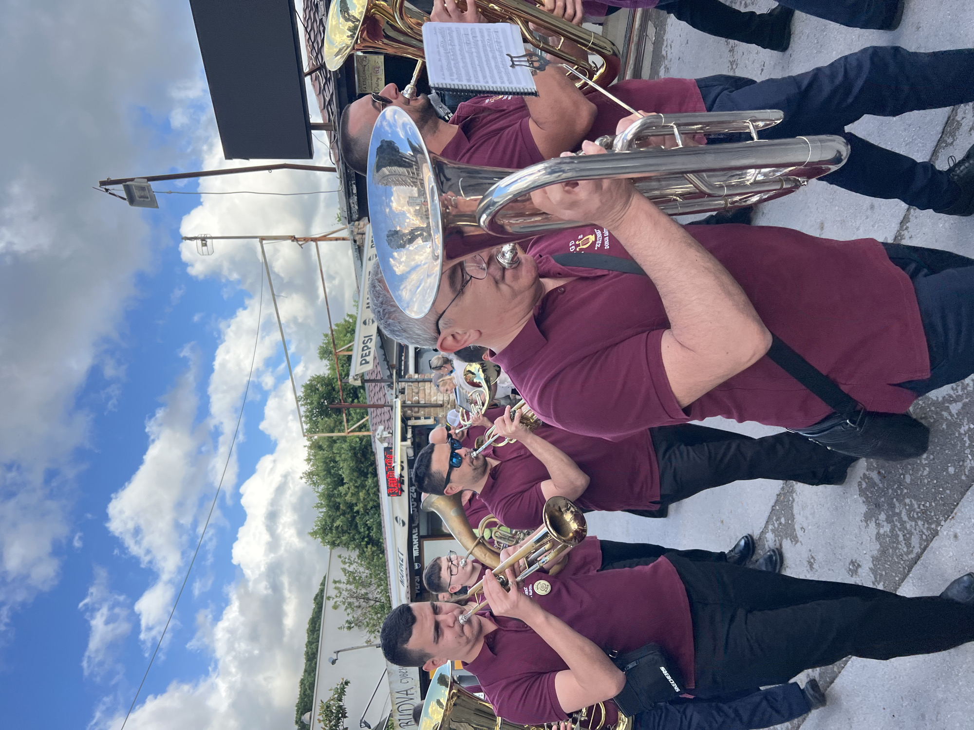 A marching band is playing in Budva, Montenegro. The musicians wear maroon shirts, black pants, and black shoes. They stand on a concrete sidewalk in front of businesses. One sign says "MARKET," another "PEPSI." A band member's bag bears the word "SKECHERS." The environment appears to be an outdoor street scene with buildings and signage visible in the background. A blue sky is visible above.