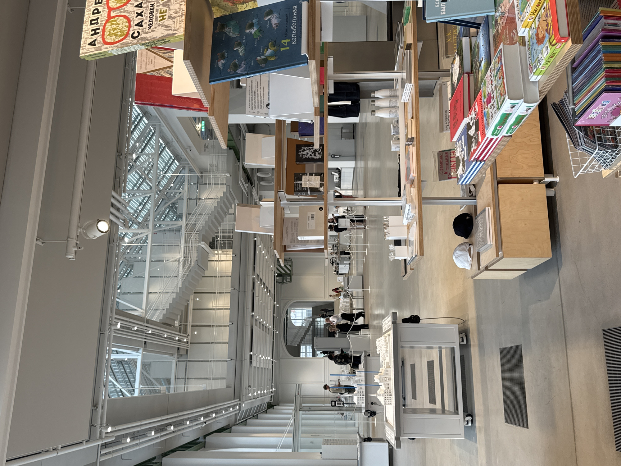 The image depicts the interior of the GES-2 House of Culture in Moscow, Russia. A multi-level space with stark white walls and modern architecture features a series of open walkways and stairs. Books are displayed at varying heights with books stacked on shelves. Some people are seated at tables. The scene appears to be a combination of a library and a public workspace.