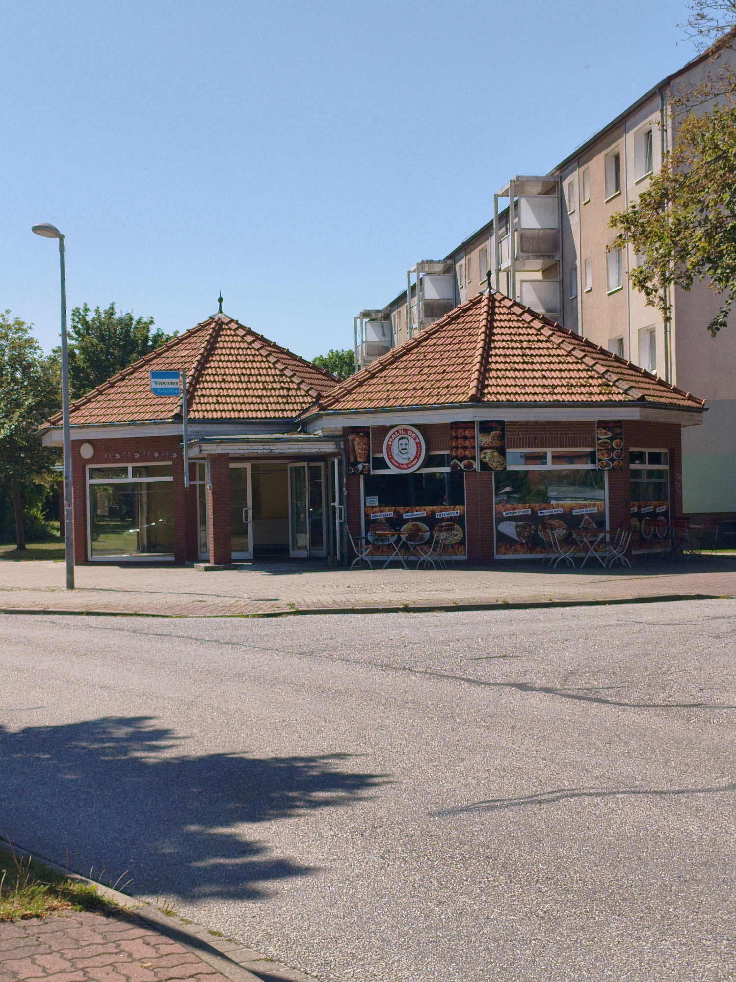 An outdoor shot shows a brick building with a red tiled roof, a restaurant named "Khalil Bey" serving Turkish food. The building is octagonal with glass windows and doors. Outside the restaurant, there are several metal tables and chairs set up for outdoor dining. The building is situated in front of a multi-story apartment building with balconies. In the foreground, there is a paved street and a sidewalk. A street lamp stands to the left of the building. The sky is clear and blue.