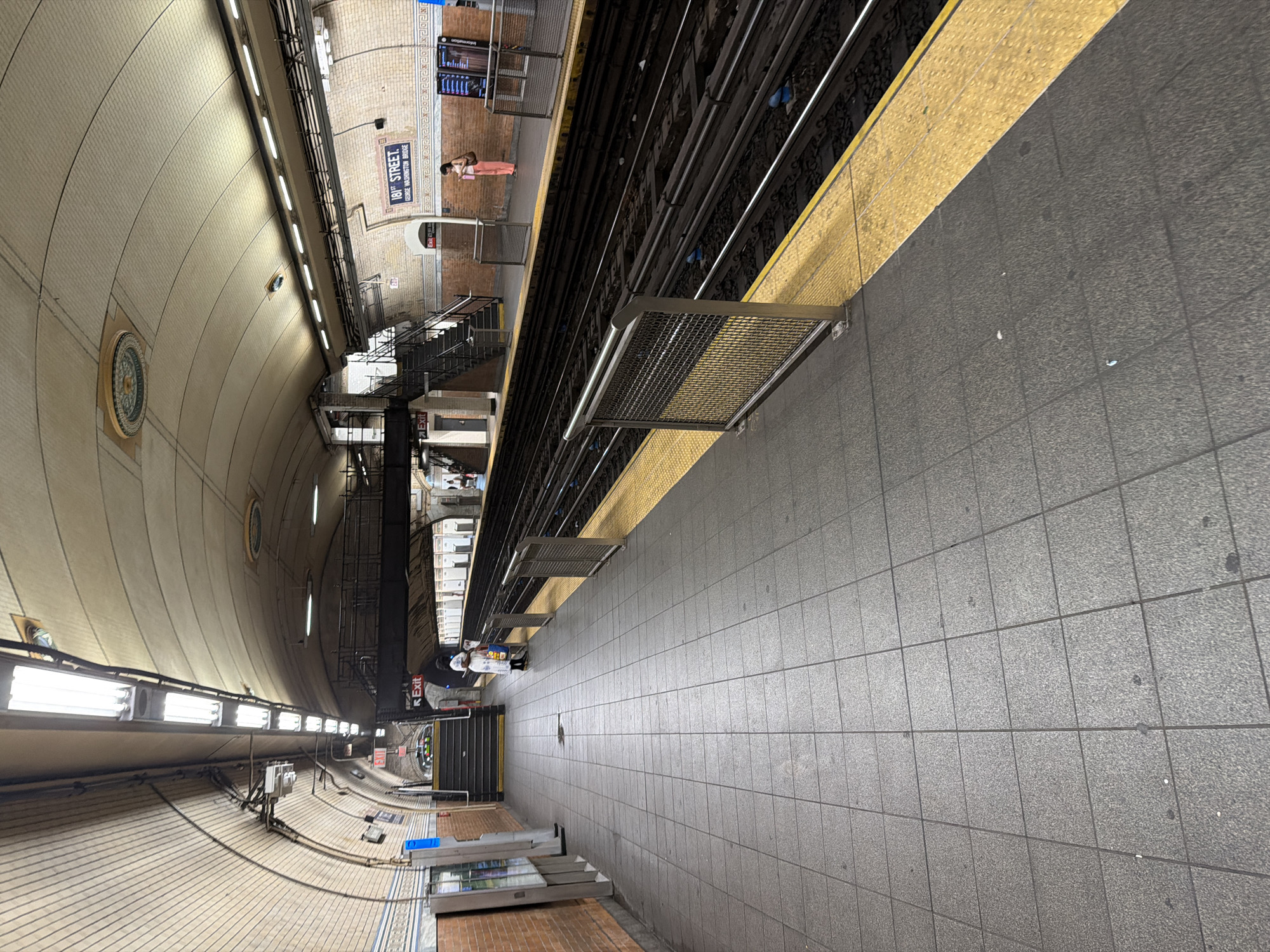 The image shows a subway station platform in New York City. The platform is tiled in gray with a yellow safety border along the track edge. Tracks are visible along the left side of the image. Along the left side of the platform is a brick wall with a sign that says "181 Street George Washington Bridge." There is a staircase leading up to the upper level of the station. There are several people on the platform. The environment is dimly lit with fluorescent lights lining the curved ceiling.