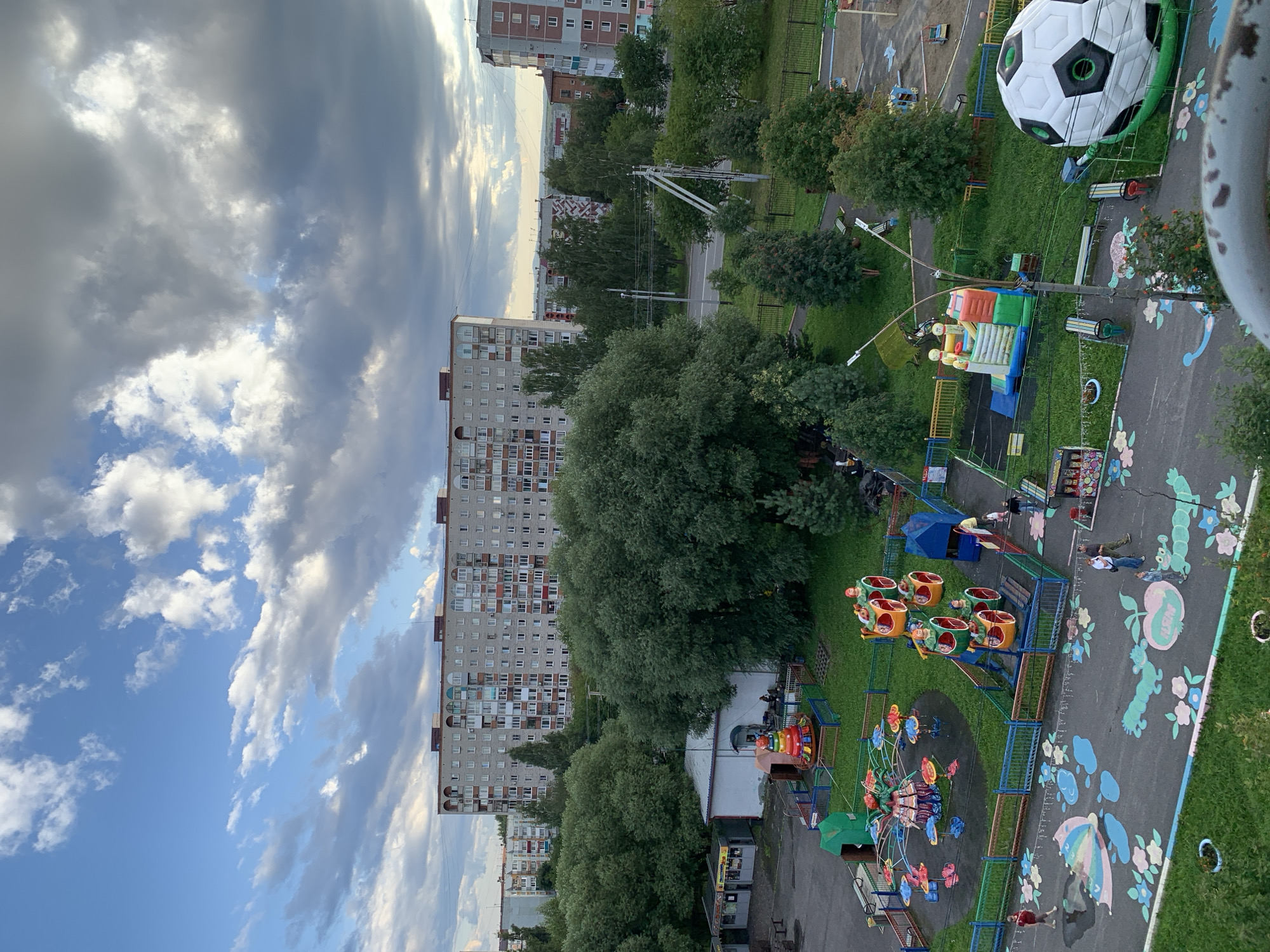An elevated, wide-angle, static shot captures a residential area in Prokopyevsk, Russia, under a partly cloudy sky. The foreground features a playground with brightly colored equipment, including a carousel, a swing set, and a bouncy castle, interspersed with grassy areas and painted pavement designs. Several people are visible, some observing the playground and others interacting with the equipment. A large inflatable soccer ball is situated to the right of the playground. Beyond the playground, mature trees form a dense green canopy, separating the play area from a road and more residential buildings in the background. These buildings are primarily multi-story apartment complexes, characteristic of Soviet-era architecture. The overall impression is one of a typical urban neighborhood with recreational facilities for children.