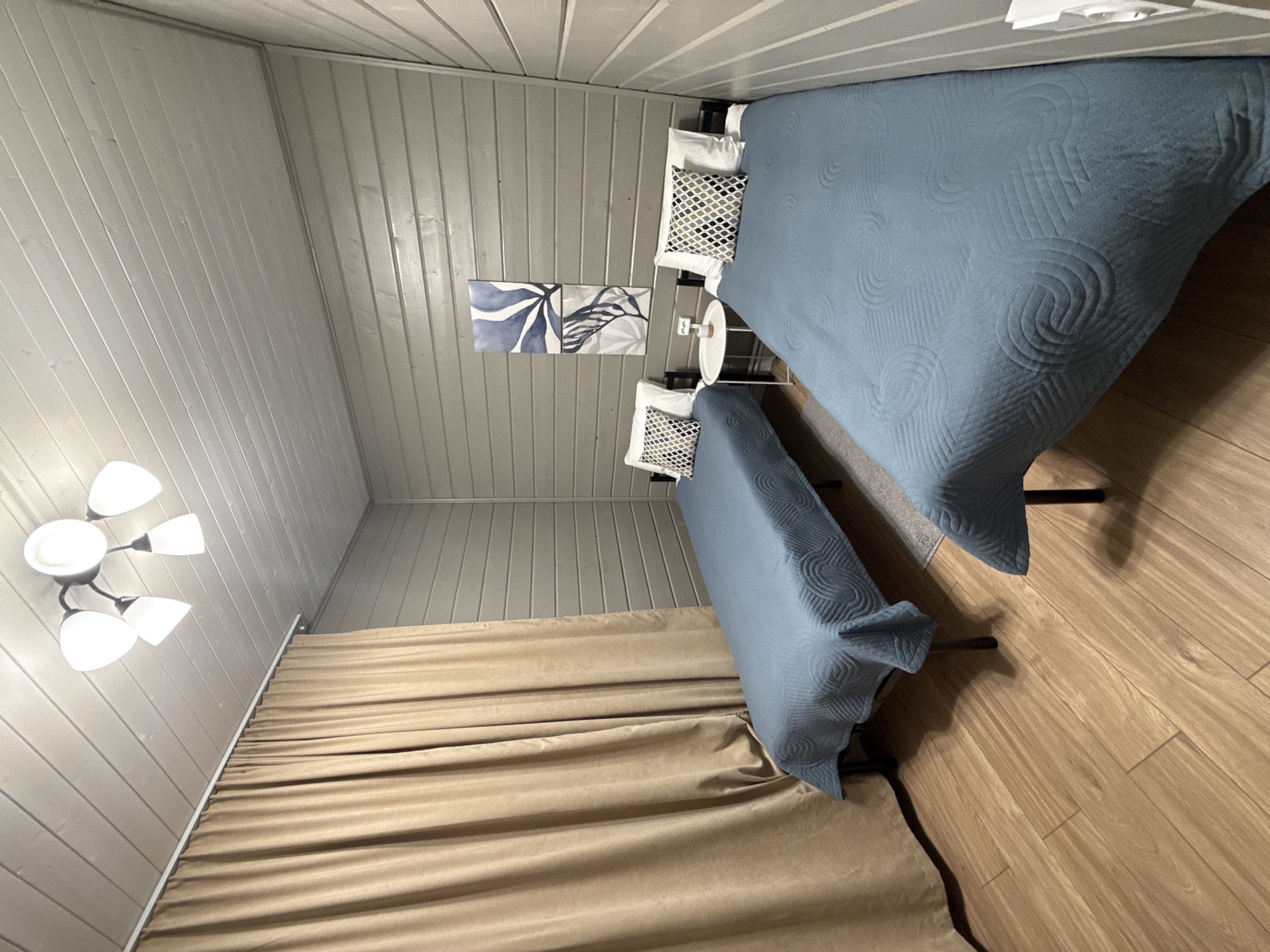 The image depicts the interior of a room, possibly in Buzhaninovo, Russia. The room features two beds covered with light blue quilted blankets. One bed is larger than the other. Each bed has white pillows with patterned pillowcases. A small round table stands between the two beds. The walls are covered with gray wood paneling. Two framed artworks depicting blue abstract floral or leaf designs are mounted on the wall above the smaller bed. A multi-bulb ceiling light fixture is visible in the lower-left corner of the image. Beige curtains drape along the wall near the smaller bed. The floor is made of light-colored wood planks.