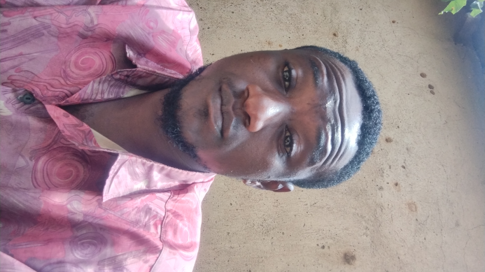 The image is a close-up selfie of a Black male against a textured, beige-colored wall. The man has short, dark, curly hair and a goatee. He is wearing a pink collared shirt with a swirling, abstract pattern in shades of pink and purple. The background wall appears to be an outdoor setting, possibly a building exterior, with some faint marks and cracks visible. The lighting suggests it is daytime. The setting is described as being in the city of Jos, Nigeria.