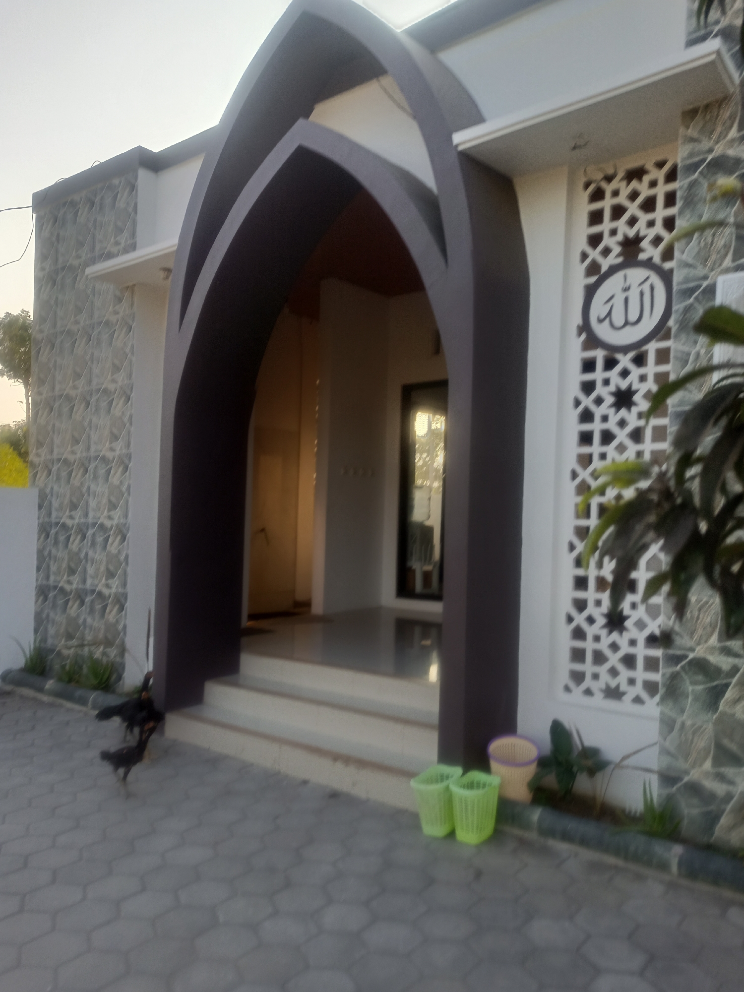 The image shows the entrance to a mosque in Nganti, Indonesia. The entrance is framed by a large, dark brown, arched structure, with the interior of the mosque visible beyond. The facade of the mosque features a decorative geometric pattern and an Arabic inscription, "Allah." To the left of the entrance, the wall is covered in textured stone-like tiles. In front of the mosque, on the paved walkway, are two green trash cans and a beige pot. Two chickens are visible on the walkway near the entrance steps, with one appearing to be pecking at the ground. The sky is overcast and appears to be late afternoon or early morning.