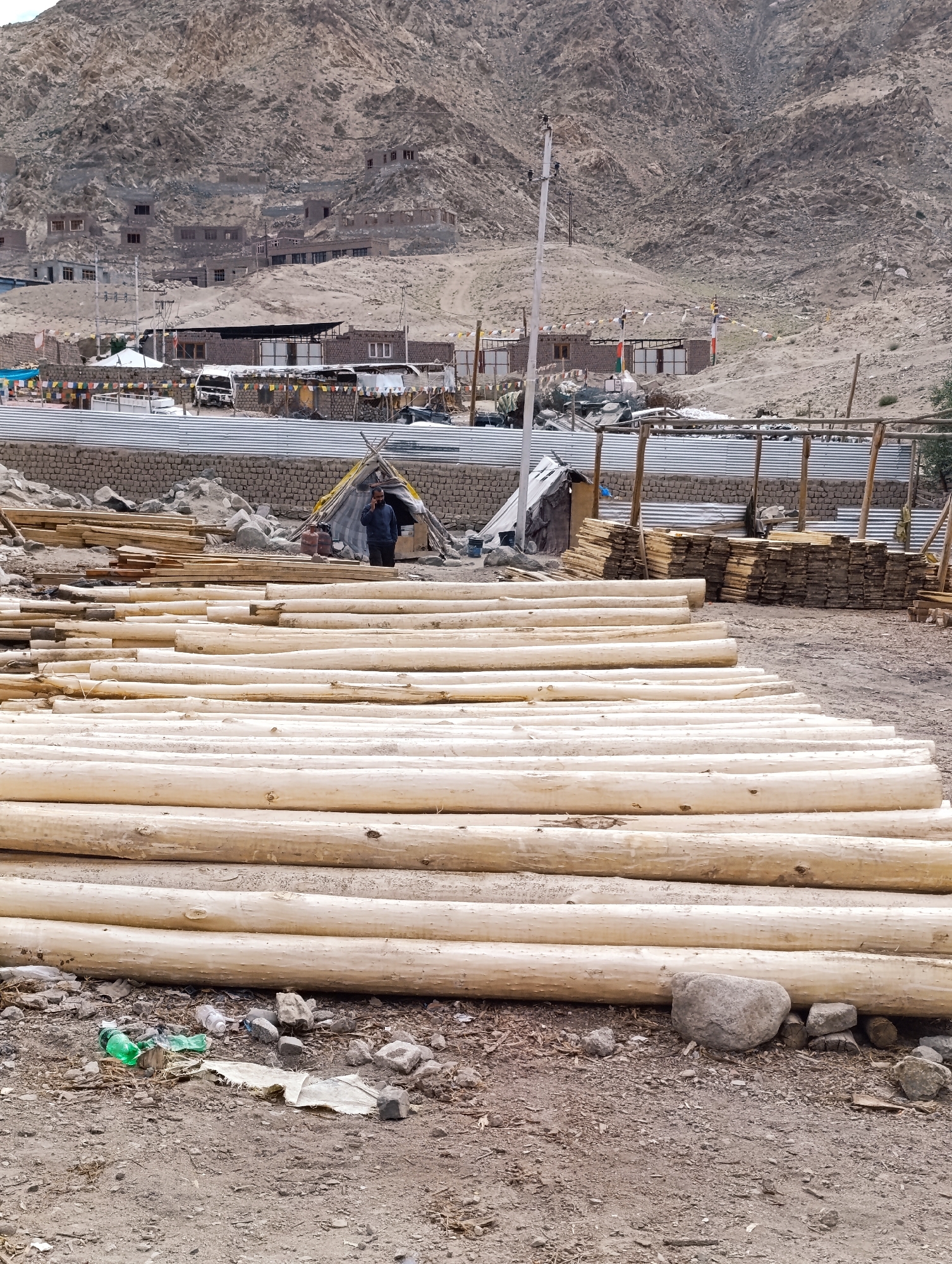 This is a photograph of a construction or lumber yard in Leh, India, situated at the base of a barren, mountainous landscape. In the foreground, a large stack of freshly cut, pale-colored logs is arranged in parallel rows, filling the lower half of the frame. The ground is composed of dirt, small rocks, and some scattered debris, including a crushed green plastic bottle and a piece of torn fabric.
In the middle ground, to the left, are piles of sawn timber. Behind these piles, a small, temporary tent-like structure is visible, with a single person standing near its entrance, appearing to be on a mobile phone. Next to the tent, two gas cylinders are present. Further behind the tent and timber piles, a long, corrugated metal wall stretches across the scene, separating the foreground and middle ground from the background. Beyond this wall, several permanent brick buildings, likely part of a settlement, are nestled into the hillside. Prayer flags, a common sight in Buddhist regions, are strung between poles in front of these buildings. In the distance, a white van is parked. The overall environment suggests a dry, arid climate typical of high-altitude desert regions. The sky is overcast, contributing to the muted color palette of the image.