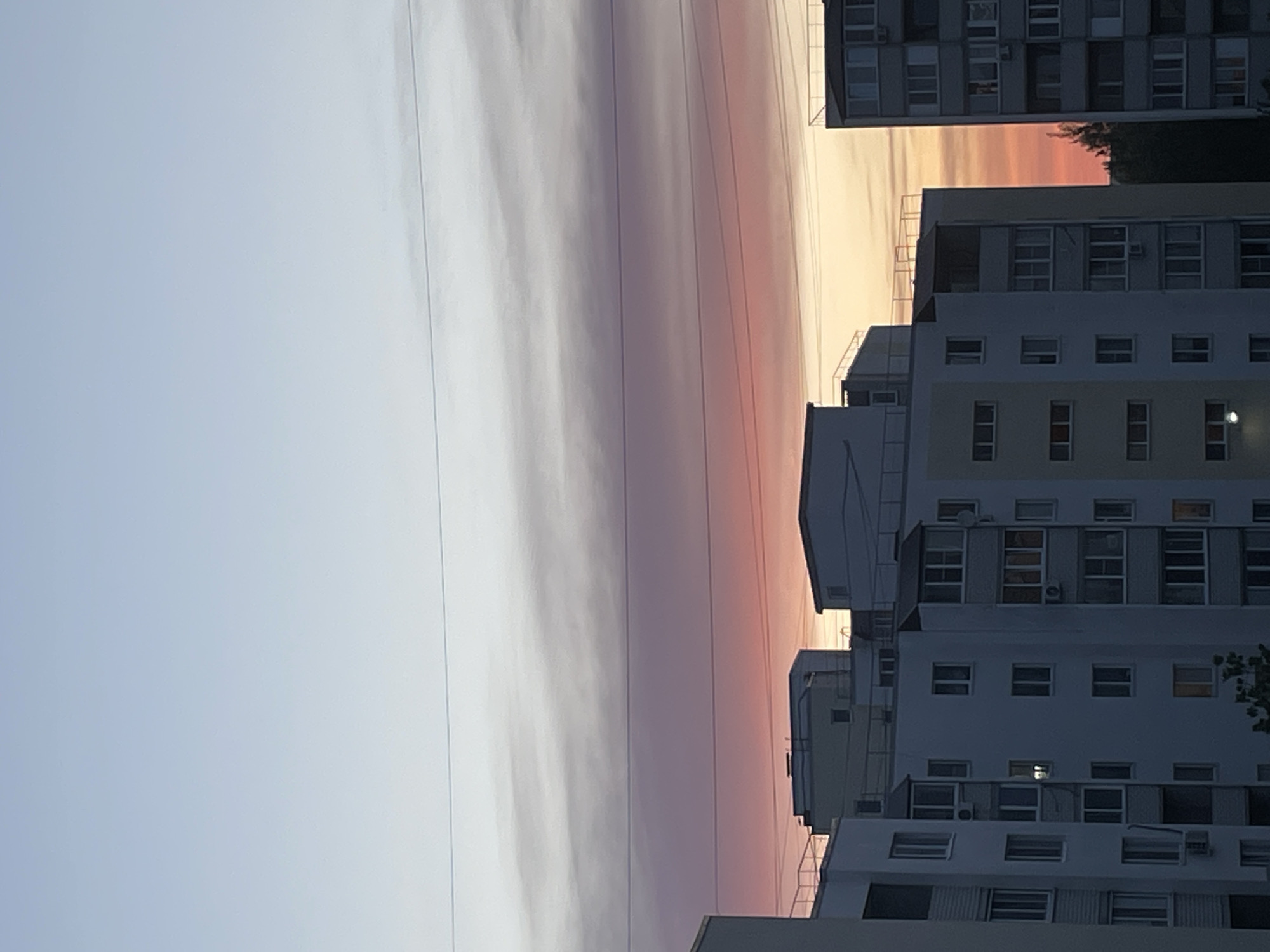 The image shows a cityscape in Saratov, Russia, at dusk or dawn. The sky transitions from a light blue to a gradient of pinks, oranges, and yellows, indicating the sun's position below the horizon. Power lines run horizontally across the sky. To the right are several multi-story apartment buildings, stacked vertically in the frame. The buildings are made of concrete and have numerous windows and balconies. Some windows are illuminated, suggesting that some residents are home. A hint of greenery can be seen to the upper right, possibly trees. The overall impression is of a quiet urban scene at the end or beginning of the day.