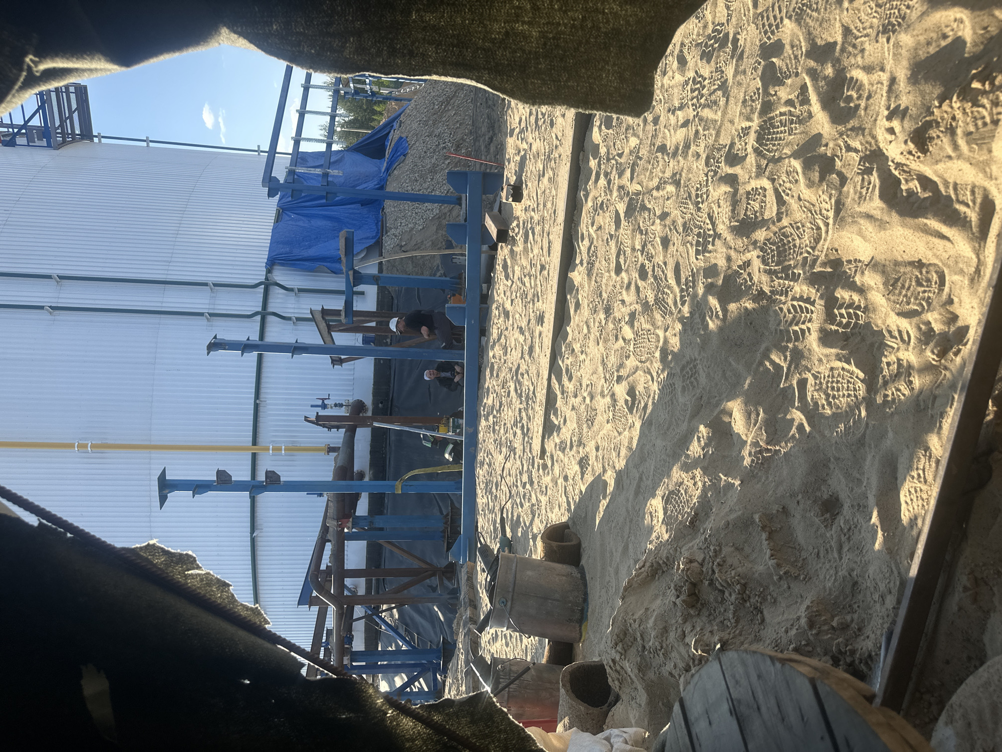 The image depicts an outdoor industrial construction site. In the foreground, sand covers the ground, marked with numerous footprints. Several buckets, one metal and one red plastic, are present, along with some cylindrical objects that appear to be concrete pipes or rolls of material. In the mid-ground, there is a blue metal structure supporting various pipes and equipment. Two individuals are visible in this area; one wearing a hard hat and dark clothing, possibly engaged in work, and another sitting, wearing a white cap and dark clothing. In the background, a large, white, cylindrical industrial tank dominates the scene, with a yellow pipe running vertically alongside it. Blue tarps are draped over a pile of gravel. The overall environment suggests ongoing construction or maintenance work at an industrial facility. The location is identified as Chikshino, Russia.