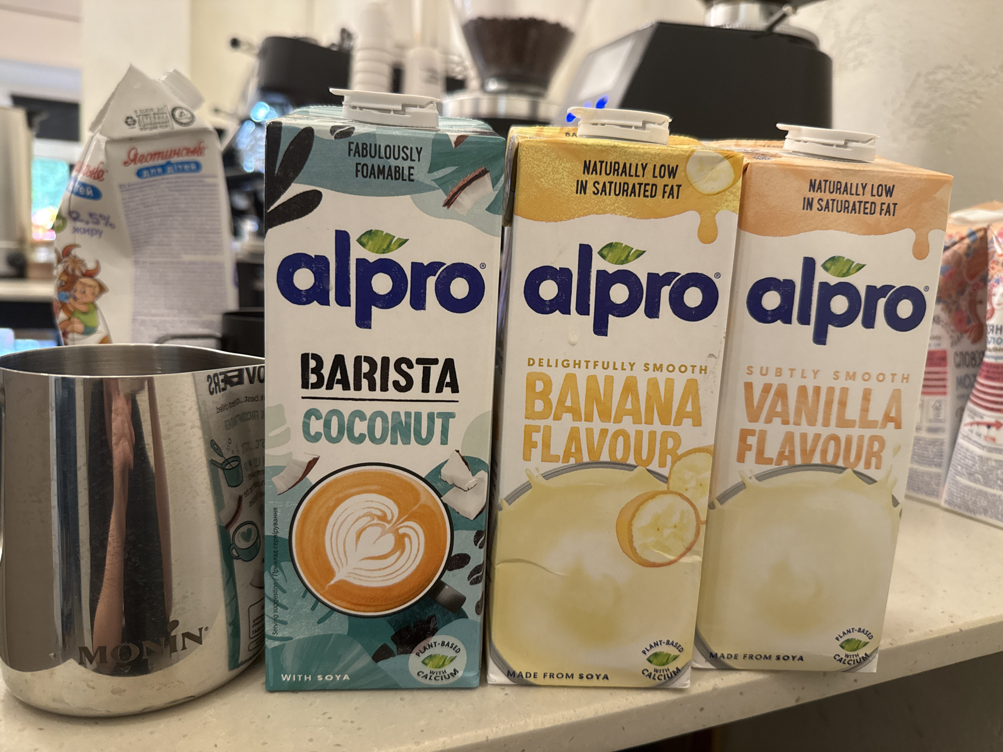 The scene, purportedly located in the city of Uzhhorod, Ukraine, depicts a close-up view of various beverage containers arranged on a light-colored, possibly speckled countertop.
In the foreground, from left to right, are three upright Alpro brand plant-based drink cartons. The leftmost carton, Alpro Barista Coconut, features a teal and white design with "FABULOUSLY FOAMABLE" printed at the top. Its lower section displays "BARISTA COCONUT" and a graphic of latte art with a heart shape. Text at the bottom indicates "PLANT-BASED WITH CALCIUM" and "WITH SOYA." Adjacent to it is the middle carton, Alpro Banana Flavour, which has a yellow and white design. It is labeled "NATURALLY LOW IN SATURATED FAT" and "DELIGHTFULLY SMOOTH BANANA FLAVOUR," accompanied by a graphic of banana slices and a splash. The text at its base reads "PLANT-BASED WITH CALCIUM" and "MADE FROM SOYA." The rightmost carton is Alpro Vanilla Flavour, with an orange and white design, also marked "NATURALLY LOW IN SATURATED FAT" and "SUBTLY SMOOTH VANILLA FLAVOUR," featuring a splash graphic. Its base also states "PLANT-BASED WITH CALCIUM" and "MADE FROM SOYA." All three Alpro cartons are sealed with white plastic screw caps; the cap on the banana carton exhibits a small blue reflective highlight.
To the far left, a polished stainless steel pitcher, bearing the brand name "MONIN" embossed near its base, stands next to the Alpro Coconut carton. Its reflective surface shows indistinct ambient light and reflections.
In the blurred background, behind the Monin pitcher and the Alpro Coconut carton, a partially crumpled white milk carton is visible. Cyrillic text "Яготинське" (Yagotynske) and "ДЛЯ ДІТЕЙ" (For Children), along with "2.5% жиру" (2.5% fat), are discernible, beside a cartoon illustration of a cow and a baby. Further back, in the upper-middle and right sections of the image, blurred components of what appears to be a coffee grinder or espresso machine are visible, including a transparent hopper filled with dark coffee beans. Additional indistinct packaging with reddish-orange patterns is partially visible on the far right background, behind the Alpro Vanilla carton. The environment appears to be an indoor setting with diffused lighting.