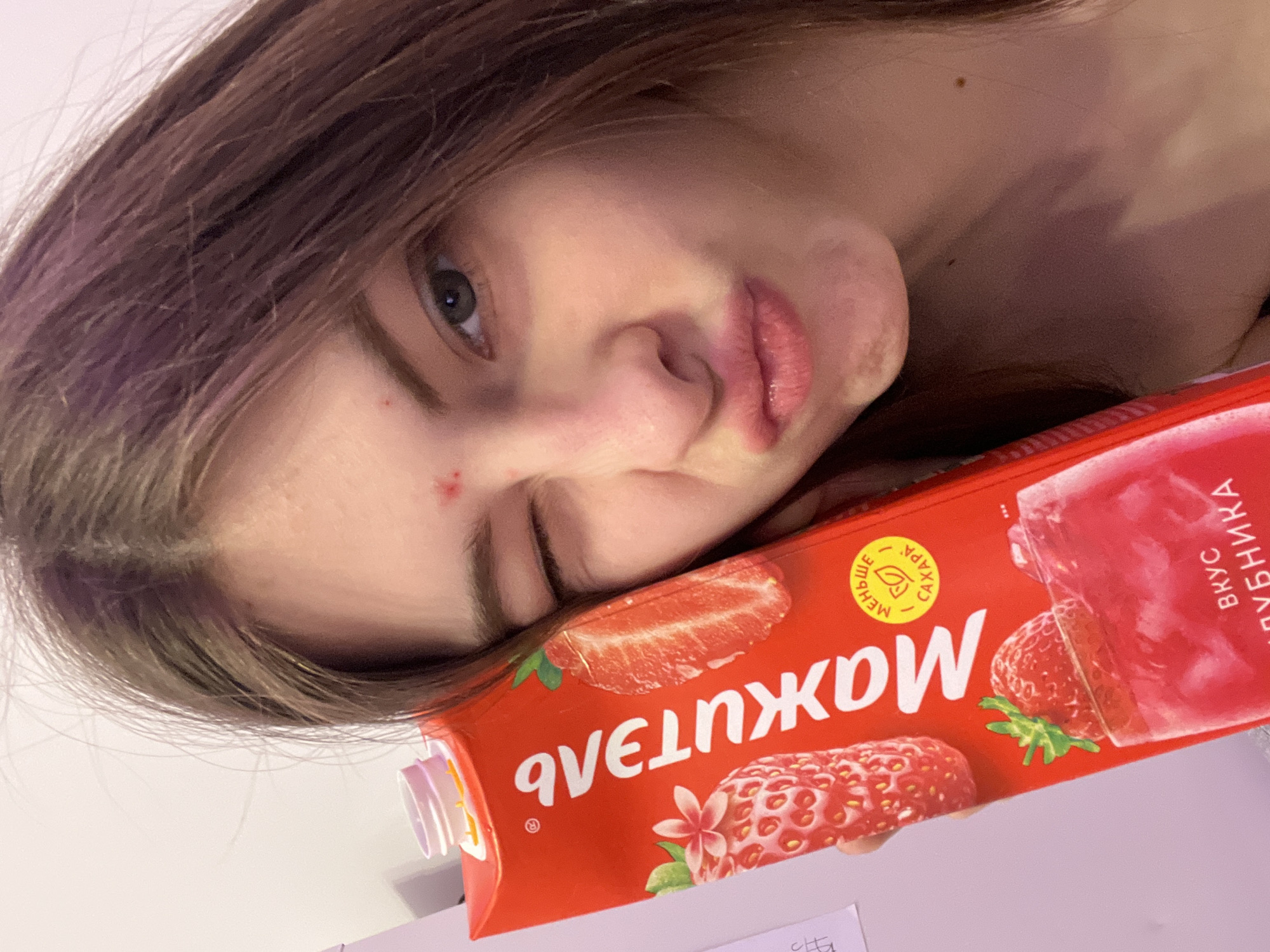 A close-up, rotated selfie features a young person, likely female, with long, dark brown hair that has a subtle purple tint. Her face is partially obscured by her hair, with one eye visible and open, looking towards the viewer, and the other eye squinted or closed. Her lips are slightly puckered, and a few red blemishes are visible on her cheek and near her nose.
She is holding a red juice carton close to her face, which is adorned with images of sliced and whole strawberries. The carton's text is in Cyrillic, aligning with the provided Saint Petersburg, Russia context. Visible text includes the brand name "МАНЖОНЖИ" (Manzhonzhi), a yellow circle indicating "МЕНЬШЕ САХАРА" (LESS SUGAR), and "ВКУС КЛУБНИКА" (STRAWBERRY FLAVOR) written vertically on the right side. The background is a plain, light-colored surface, suggesting an indoor setting. The overall mood is casual and playful.